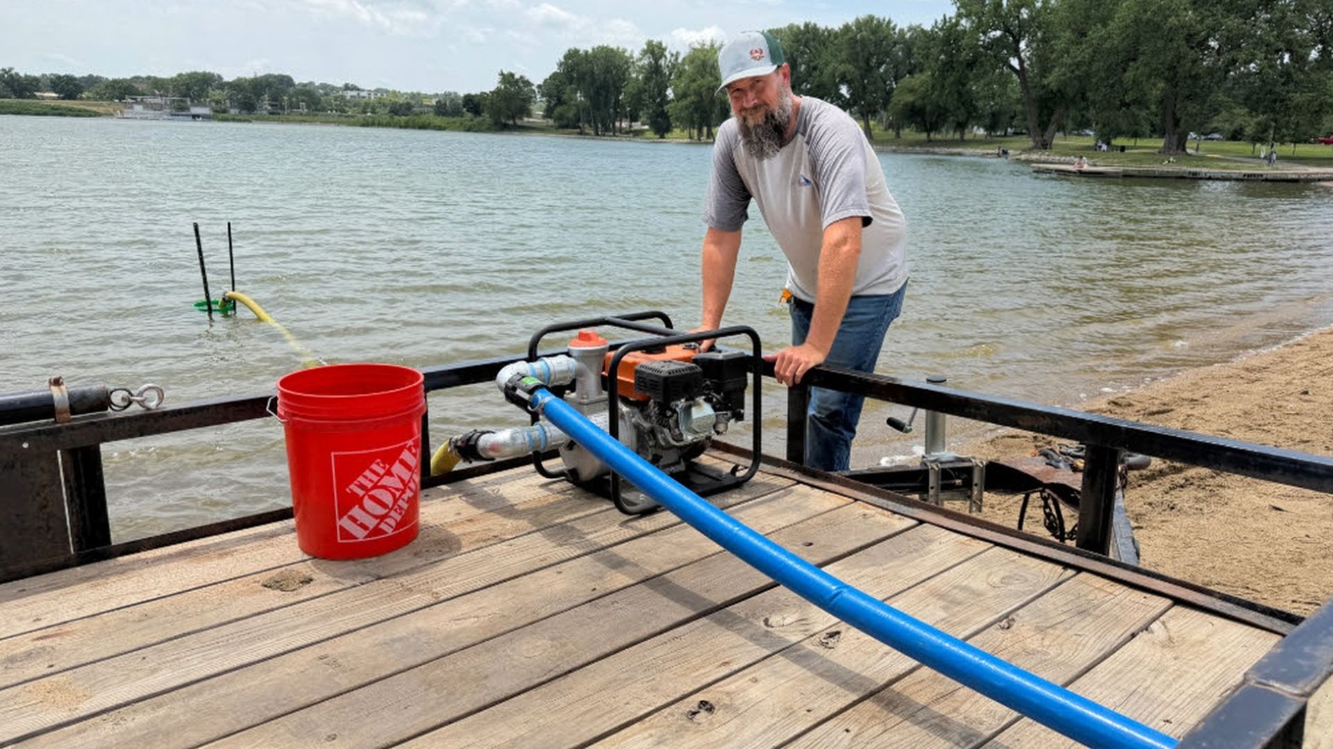 Bearded man in a gray T-shirt and cap stands on a wooden dock by a lake, operating a portable gas pump with a blue hose. A red Home Depot bucket sits nearby; trees line the shore.
