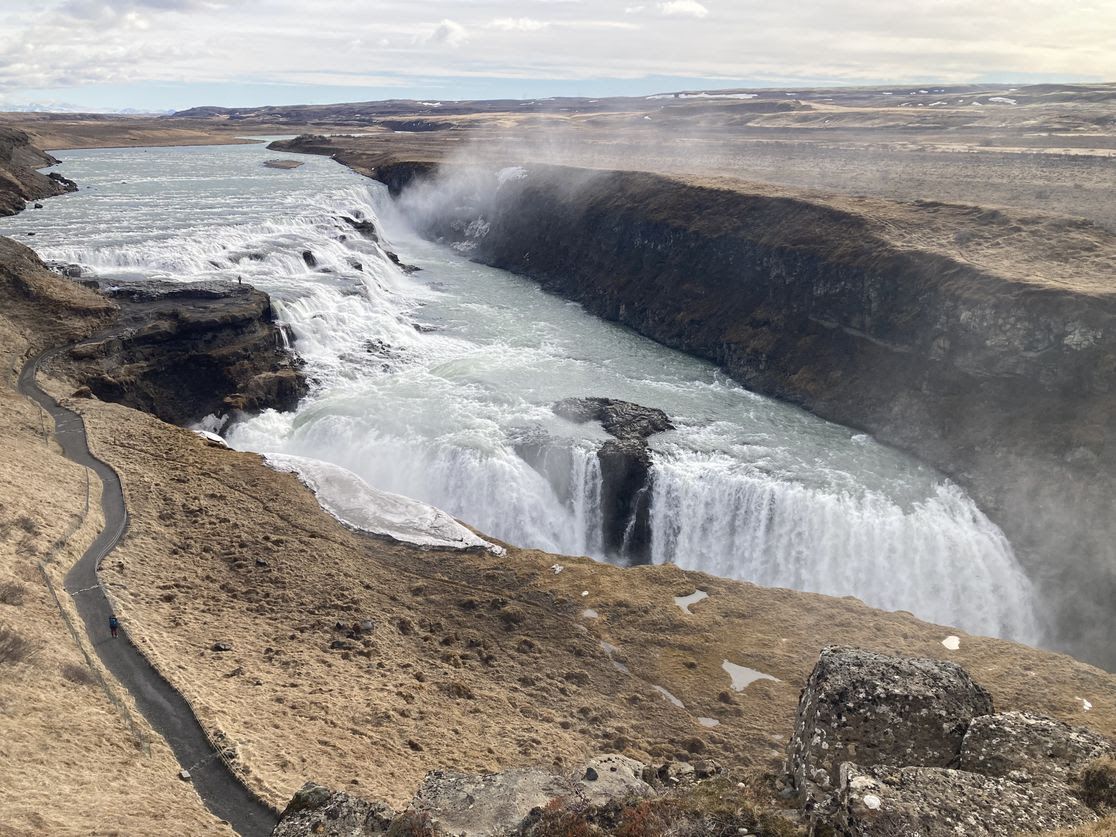 Wide waterfall plunges into a deep canyon with mist rising from the spray. A winding path runs along the left edge where a lone hiker walks, flanked by brown grasses and rocky foreground.