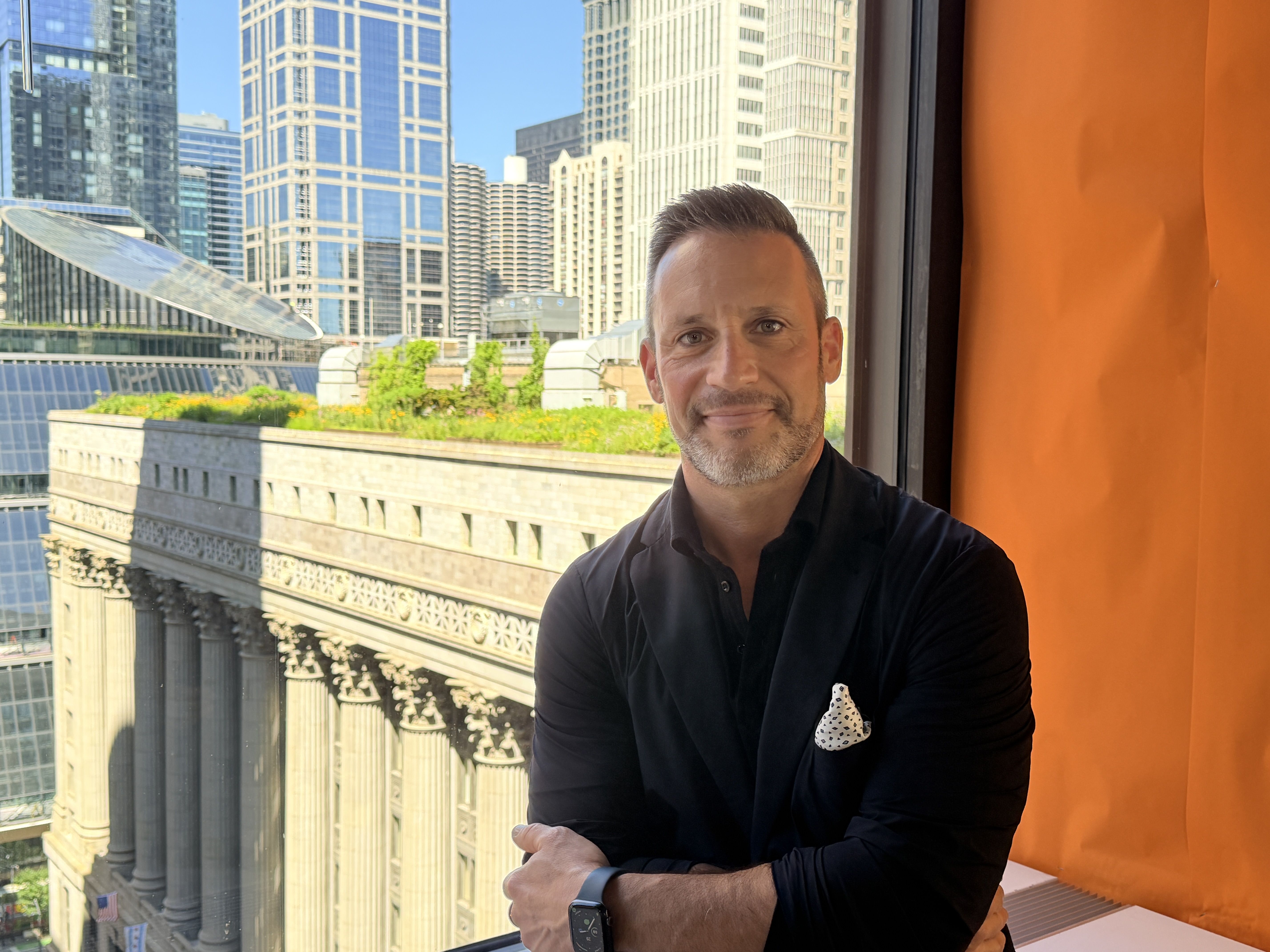 Smiling man in a black shirt with a white polka-dot pocket square leans on a window frame. Behind him, a city of tall blue glass skyscrapers under a clear sky, with an orange curtain to the right.