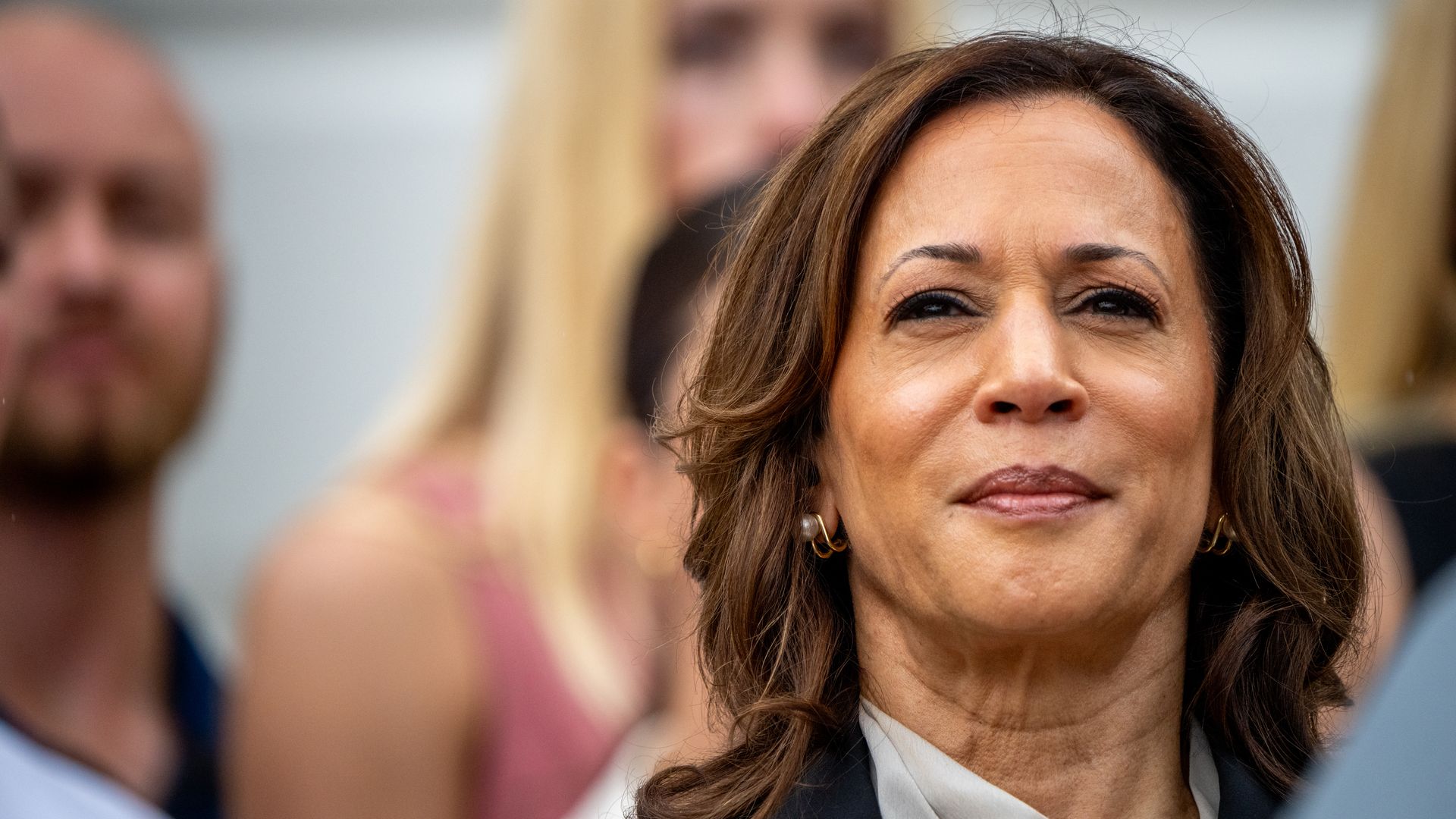 Close-up of Kamala Harris wearing pearl earrings and black jacket, smiling slightly, with blurred people in the background.