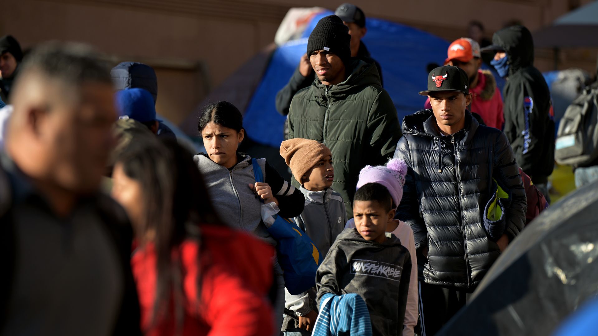 A photo of a line of migrants of differing ages, including children and adults, standing outside in winter clothing.