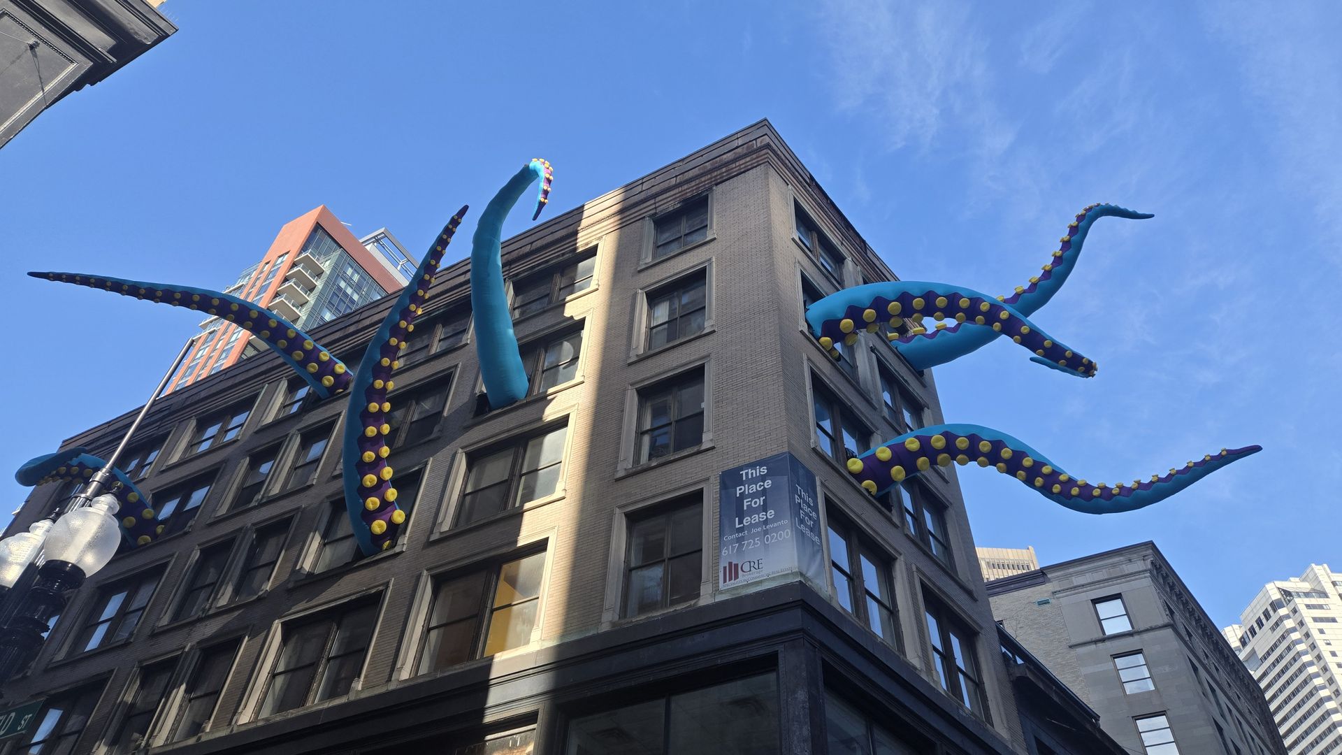 Large blue and purple tentacles with yellow suction cups emerge from windows of a beige multi-story building under a clear blue sky in an urban area.