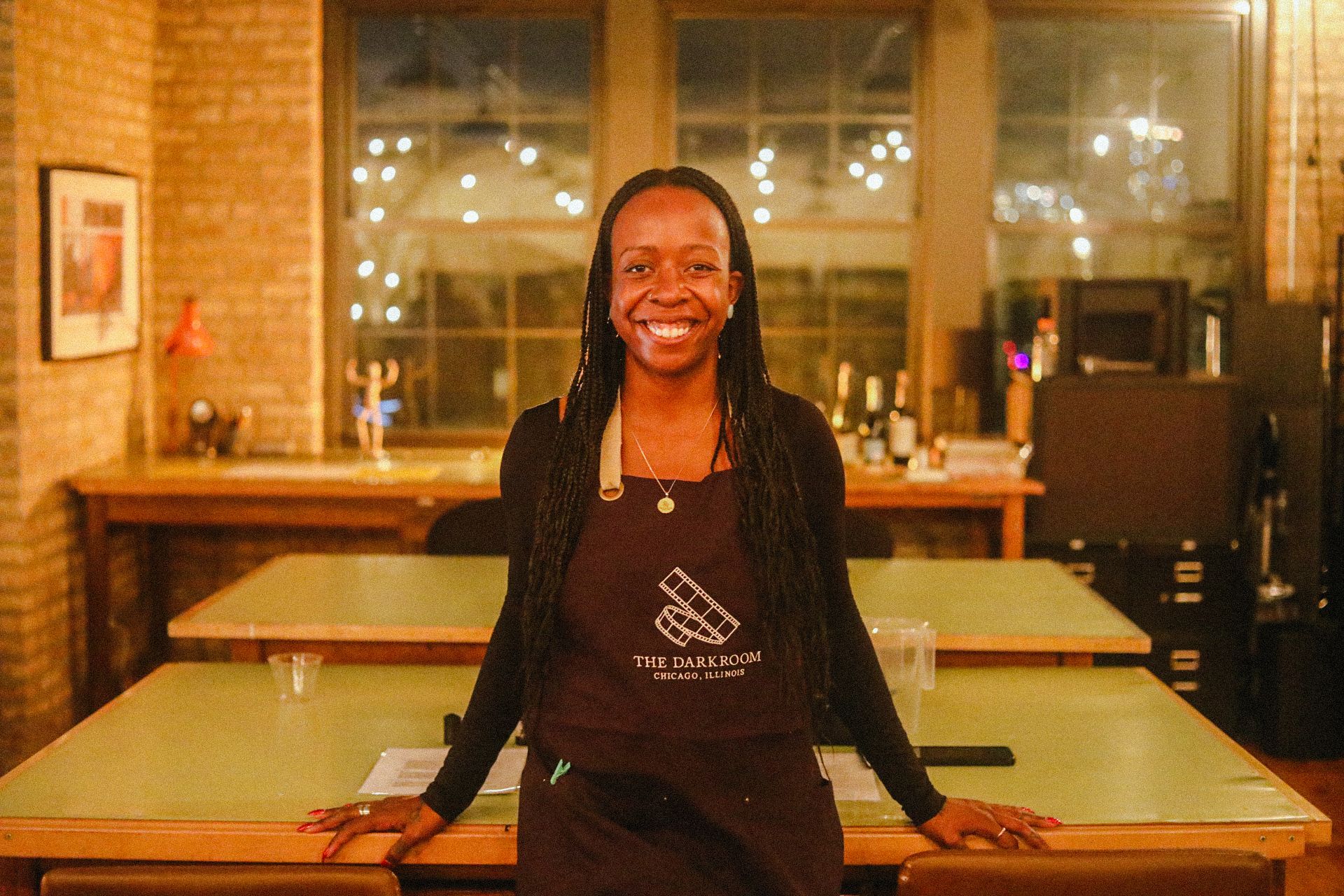 Rosondunnii Marshall wearing a "The Darkroom Chicago, Illinois" apron, standing in a warmly lit room with wooden tables and brick walls, festive string lights in the background.