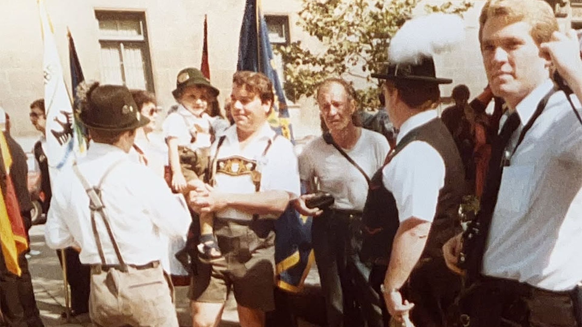 Group of men in traditional Bavarian attire (lederhosen, hats, knee socks) stand on a sunny city street during a parade; flags wave nearby as one man holds a small child.