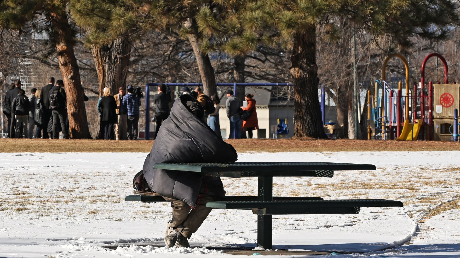 Snowy park scene with a dense canopy of evergreen trees; a group of people in jackets near a colorful playground on the right; a person in a dark coat sits at a green picnic table in the foreground.