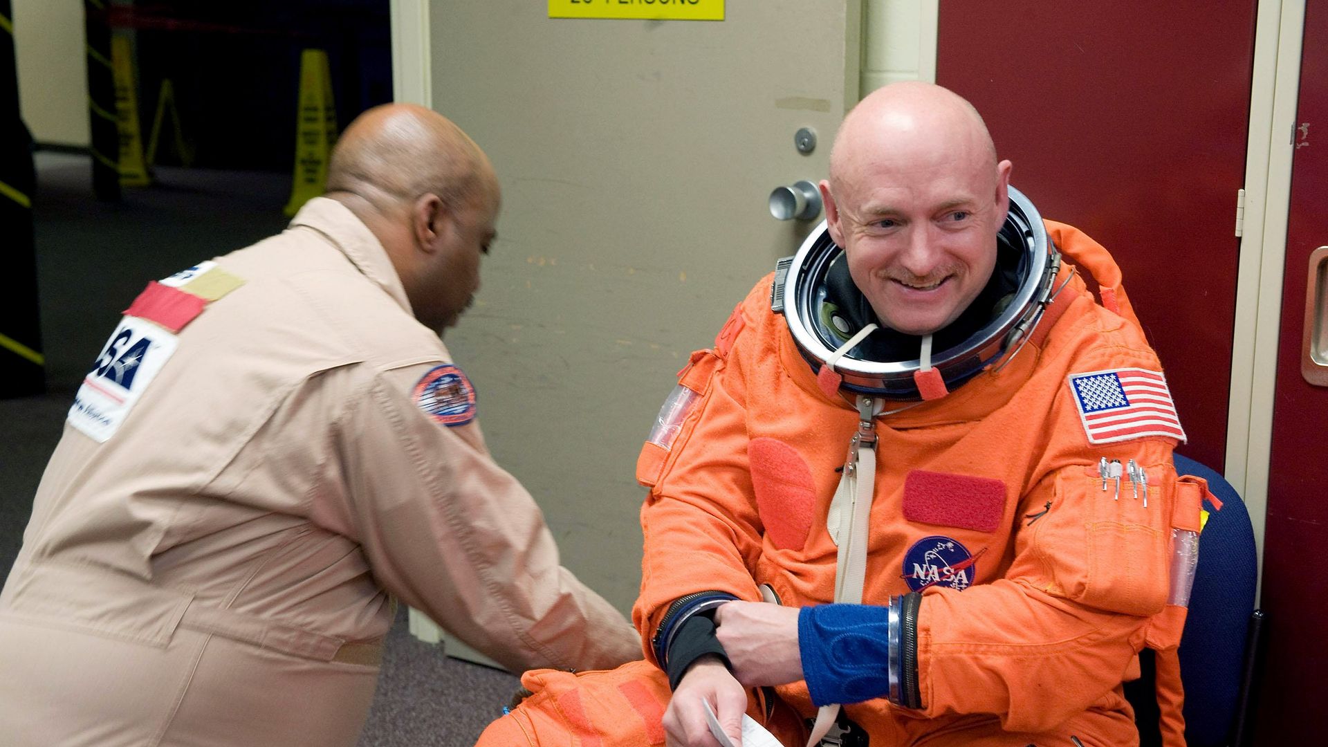Then-astronaut Mark Kelly, dressed in an orange space suit, is shown with a NASA worker dressed in tan.