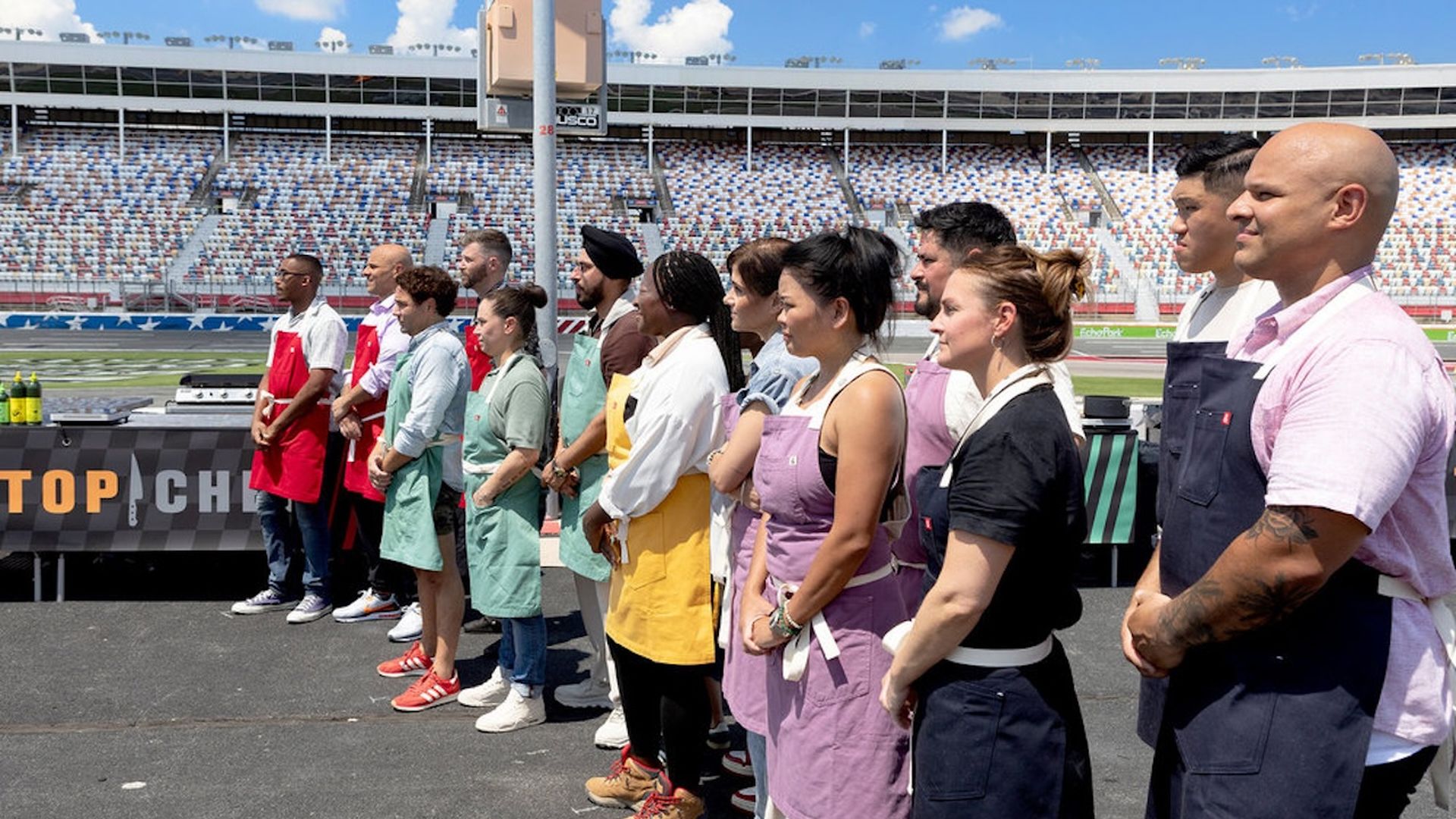Group of chefs and contestants wearing colorful aprons standing in a line on a racetrack under blue sky with clouds, preparing for a cooking challenge event.
