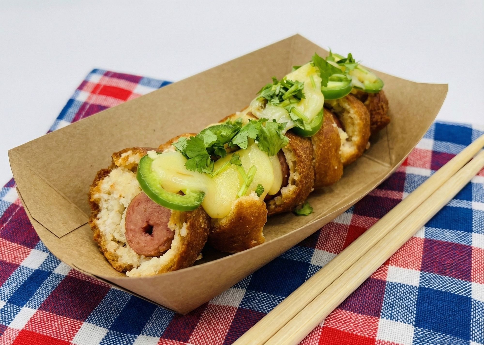 Sliced fried snack with sausage inside, topped with melted cheese, jalapeño slices, and fresh cilantro, served in a cardboard tray on a red, white, and blue checkered cloth with wooden chopsticks.