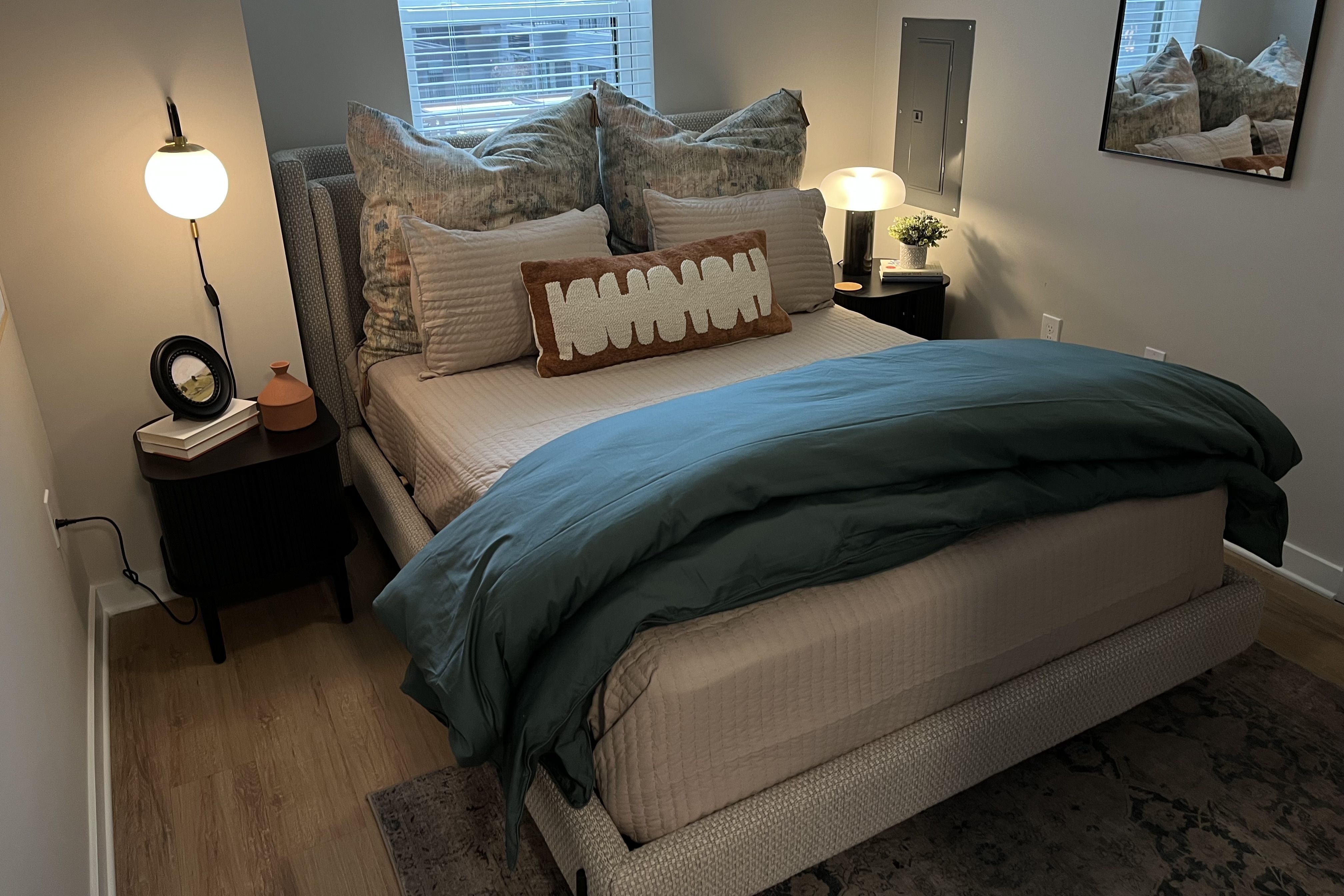 Cozy bed with beige bedding, teal comforter, and multiple pillows including a brown decorative one, flanked by black nightstands with lamps, a mirror, and window blinds in a modern bedroom.