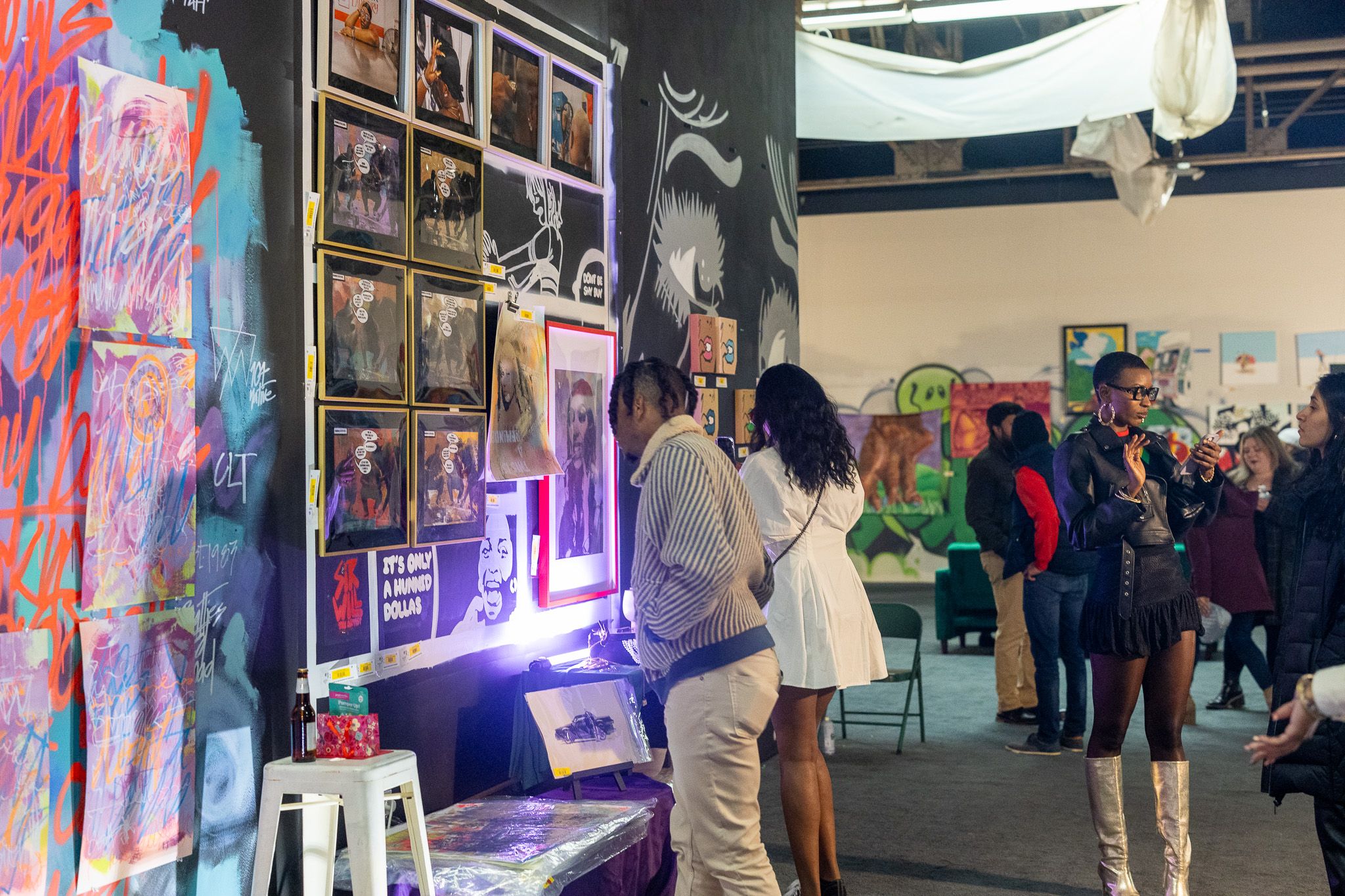People viewing diverse artwork in an urban gallery with graffiti-style walls, framed photos, colorful paintings, and a mix of casual and stylish attire in a vibrant indoor setting.