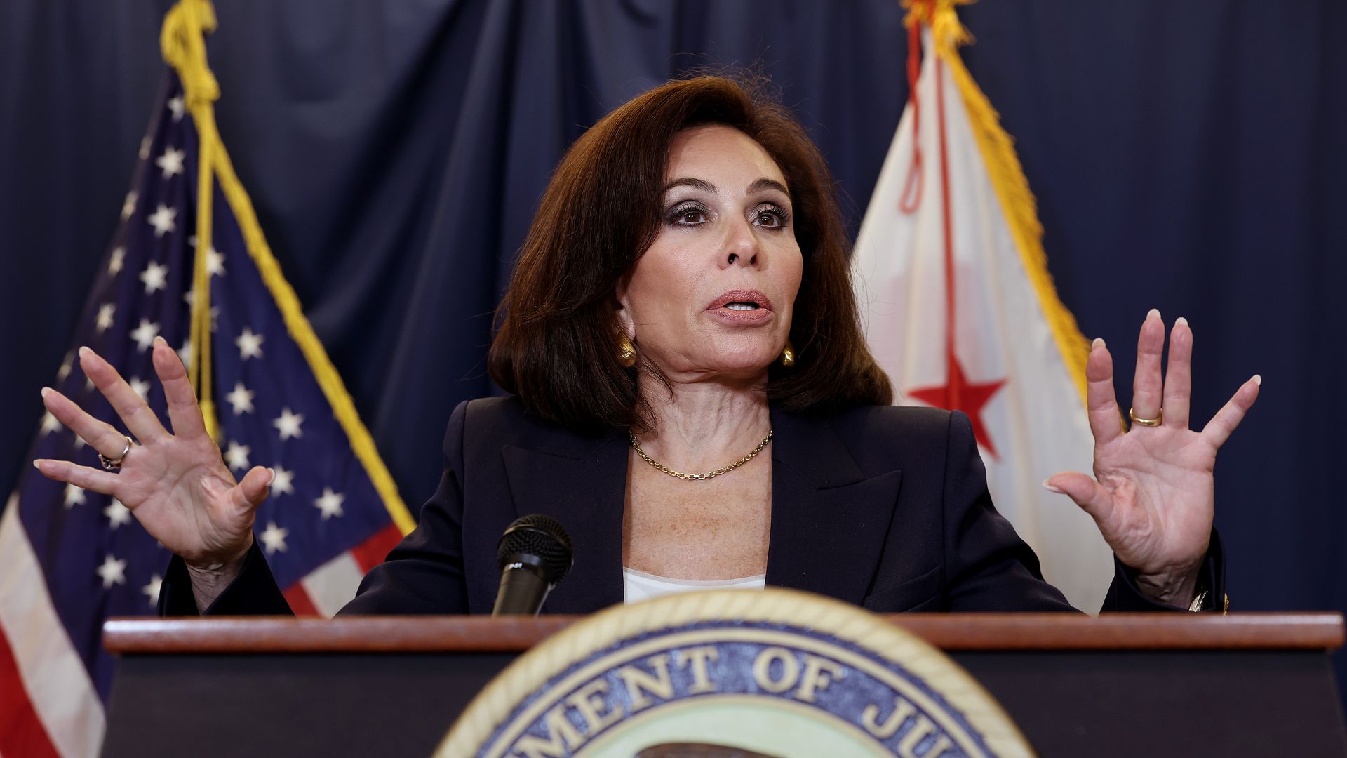 Woman with dark hair in a black blazer speaking at a podium with the Department of Justice seal, with American and California flags in the background.