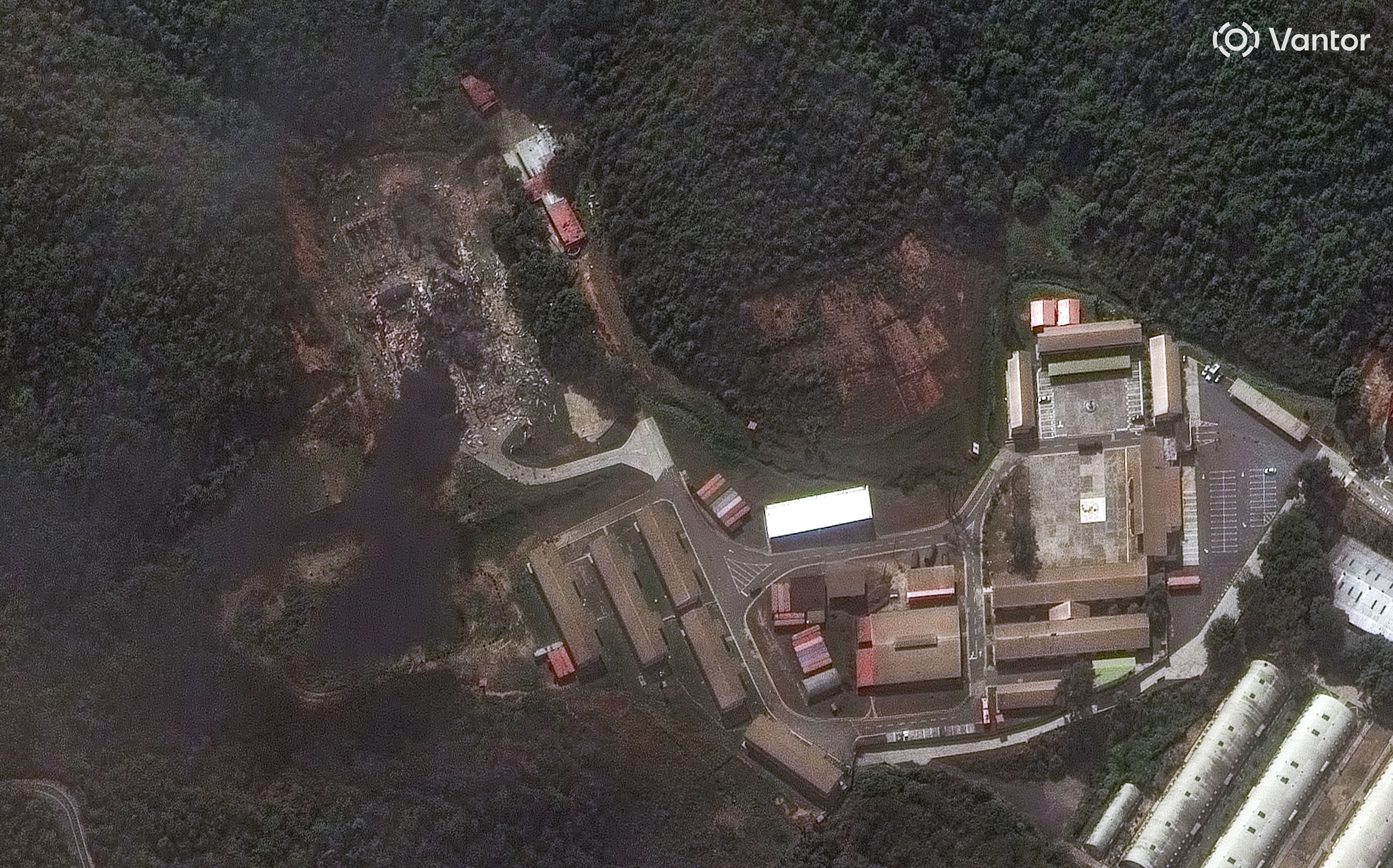 Aerial view of a compound with multiple buildings, roads, and parking areas surrounded by dense green forest. Some sections show cleared or damaged land.