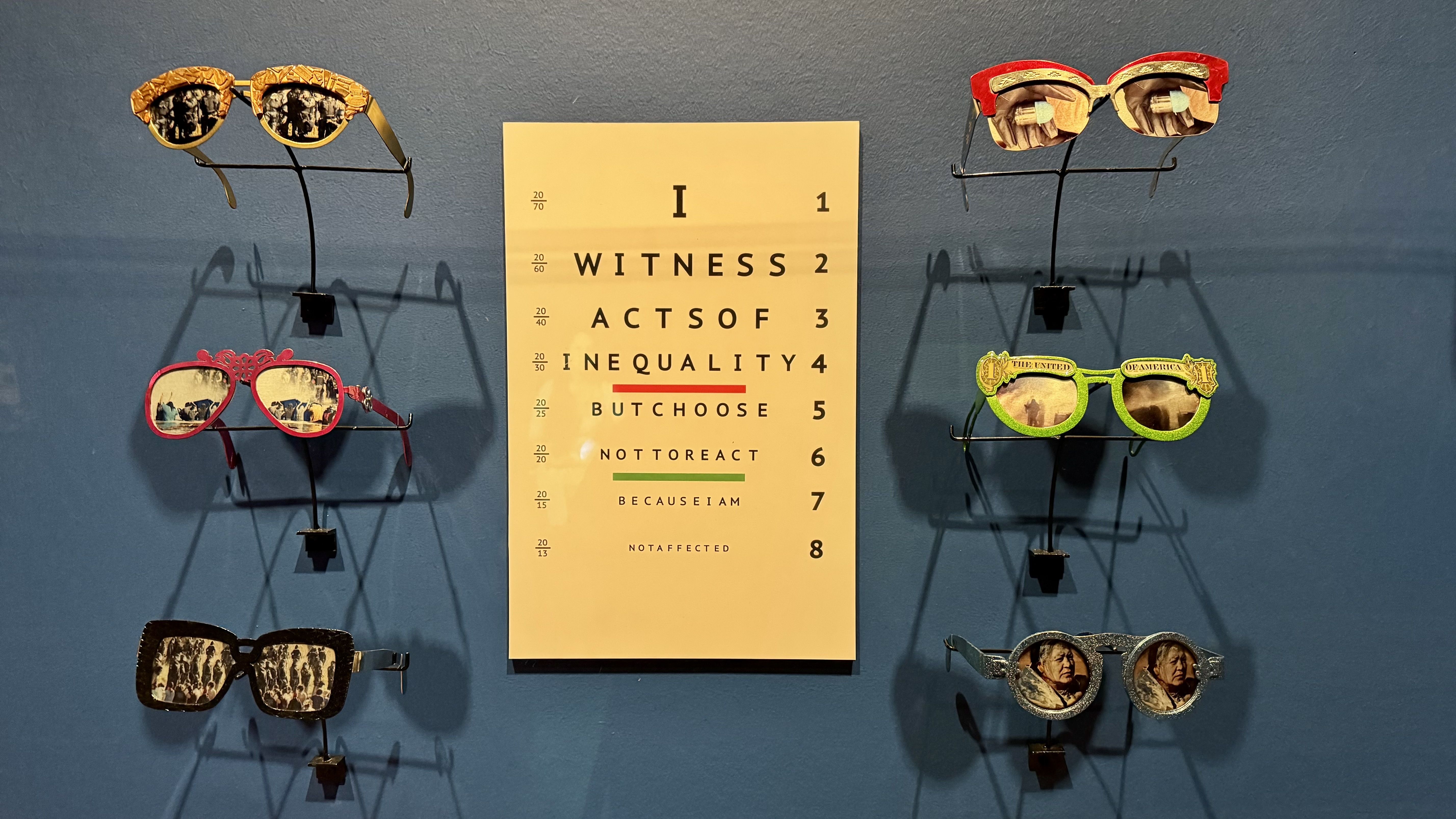 Eyeglasses with vintage black-and-white photos on lenses arranged on a blue wall around a beige eye chart reading "I WITNESS ACTS OF INEQUALITY BUT CHOOSE NOT TO REACT BECAUSE I AM NOT AFFECTED".