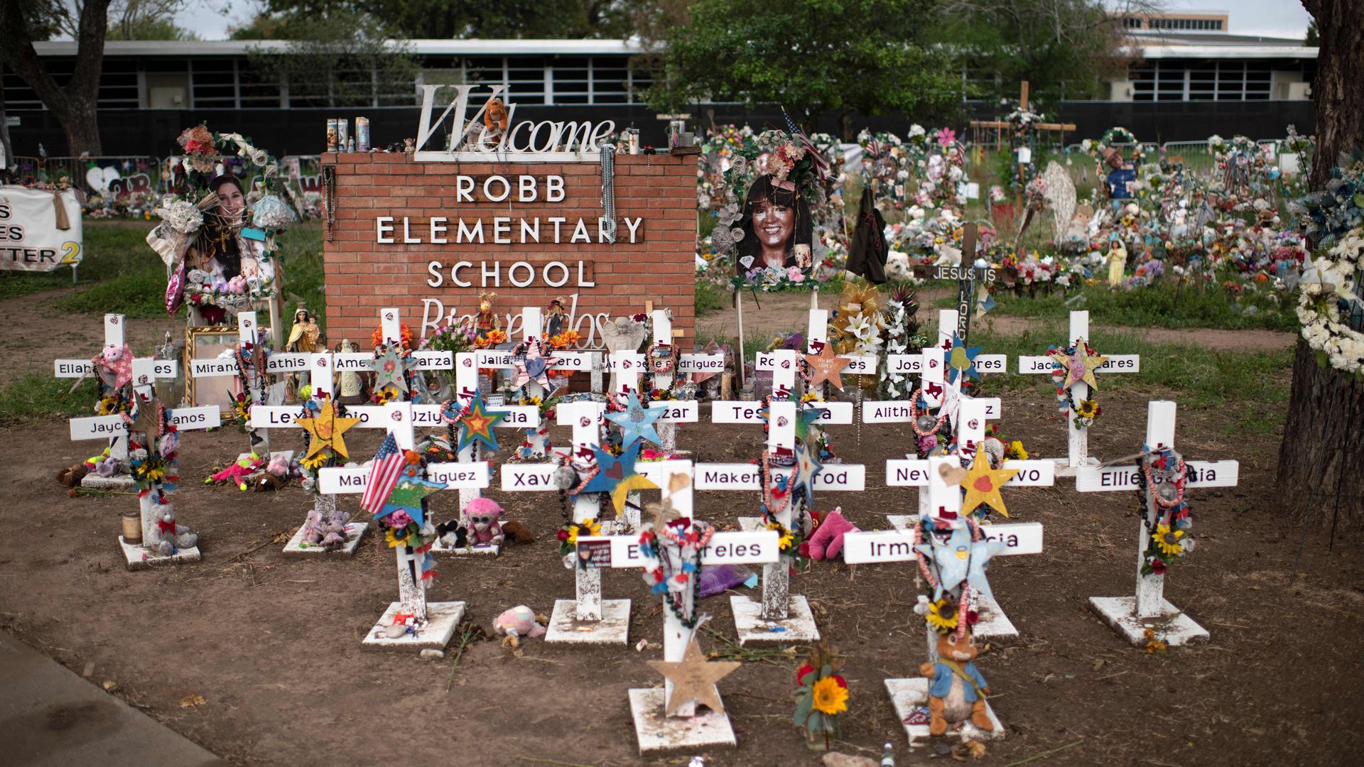 Photo of crosses bearing flowers and names standing in the ground in front of the Robb Elementary School sign
