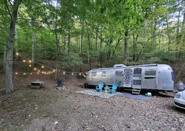 An Airstream sits on a camp site with string lights and lawn chairs.