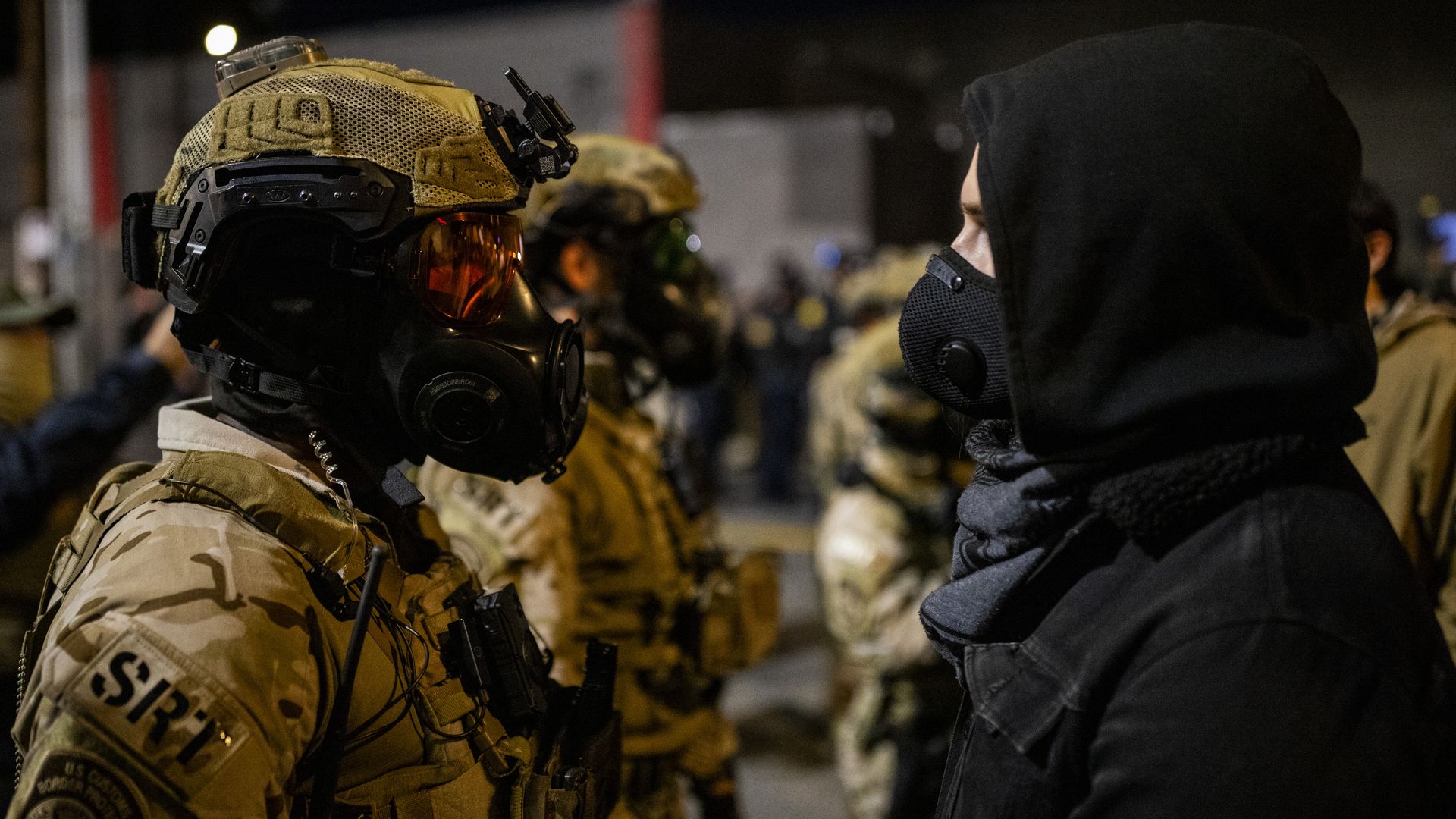 A soldier in tan camouflage gear and gas mask faces a person in a black hooded jacket and black mask at night with more soldiers blurred in the background.