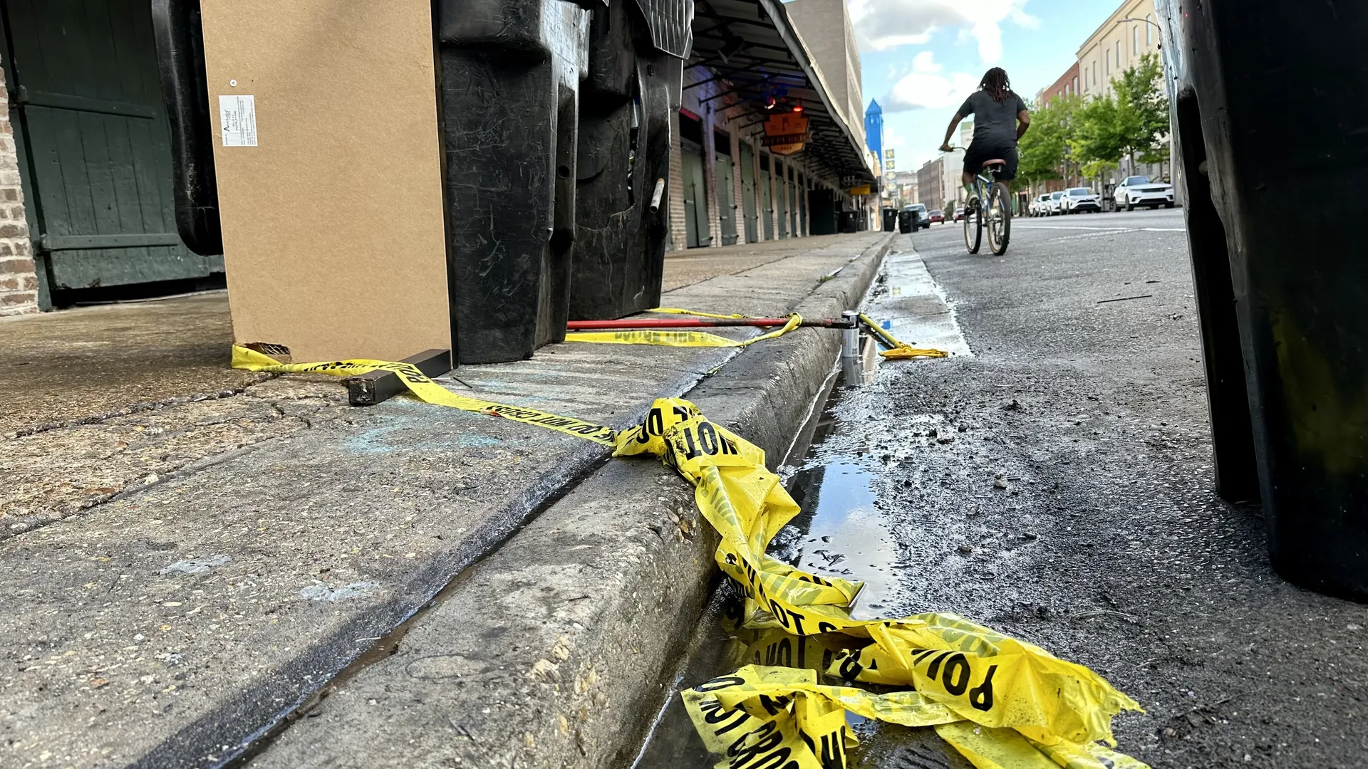 Low-angle city street scene: cardboard box and large black dumpsters by the curb, yellow caution tape, a cyclist riding away on wet pavement, storefronts and trees in the background.