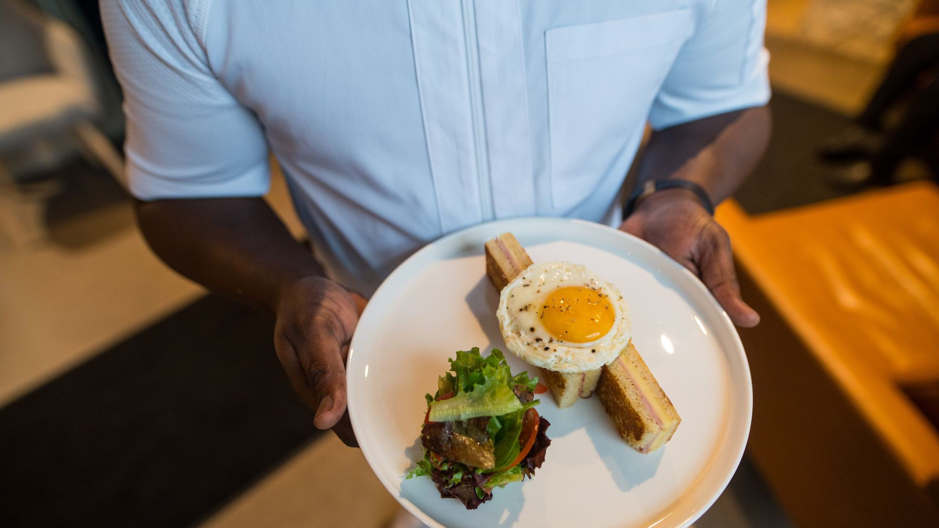 Person in a light blue shirt holds a white plate featuring a sunny-side-up egg on top of a crosswise stack of toasted sandwiches, with a small mixed greens salad on the side.