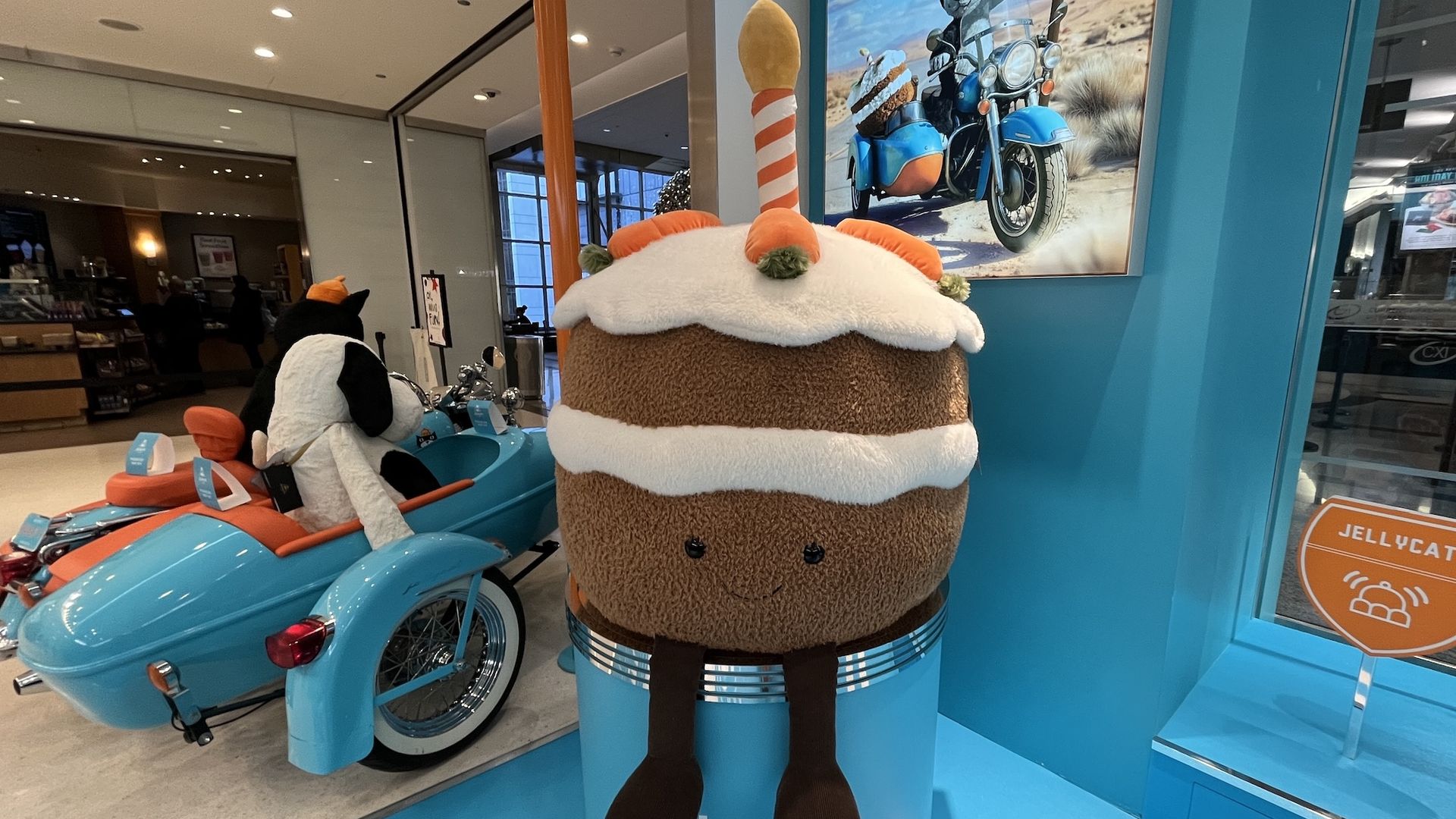 Large plush cake with white frosting, orange decorations, a striped candle, and smiling face next to a blue motorcycle with a black and white dog plush in sidecar, inside a store.