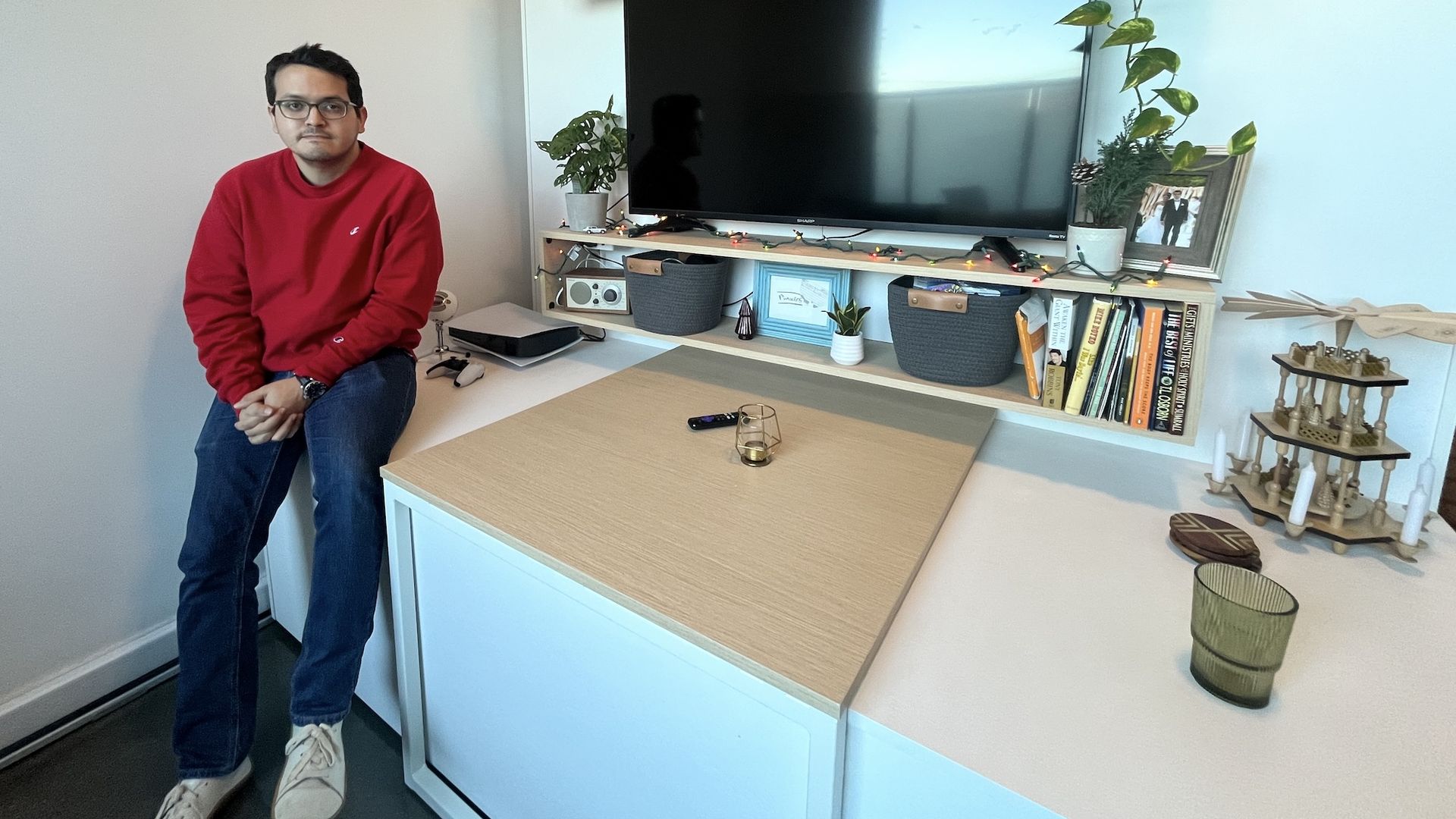 Man in red sweatshirt and jeans sits on counter next to a table and in front of a TV and entertainment center.