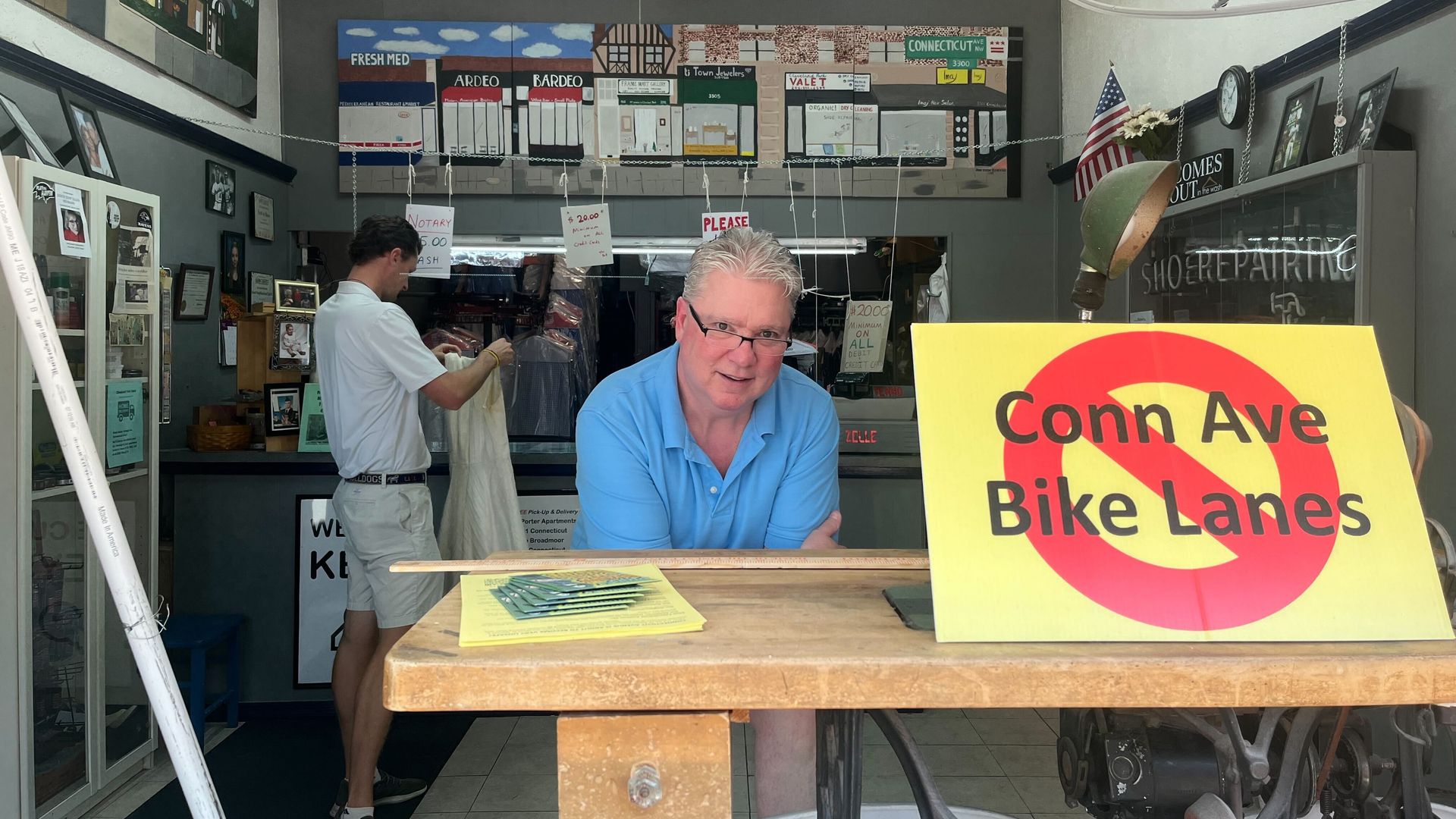 Dry cleaner shop owner poses next to anti "Conn Ave Bike Lanes" sign on storefront