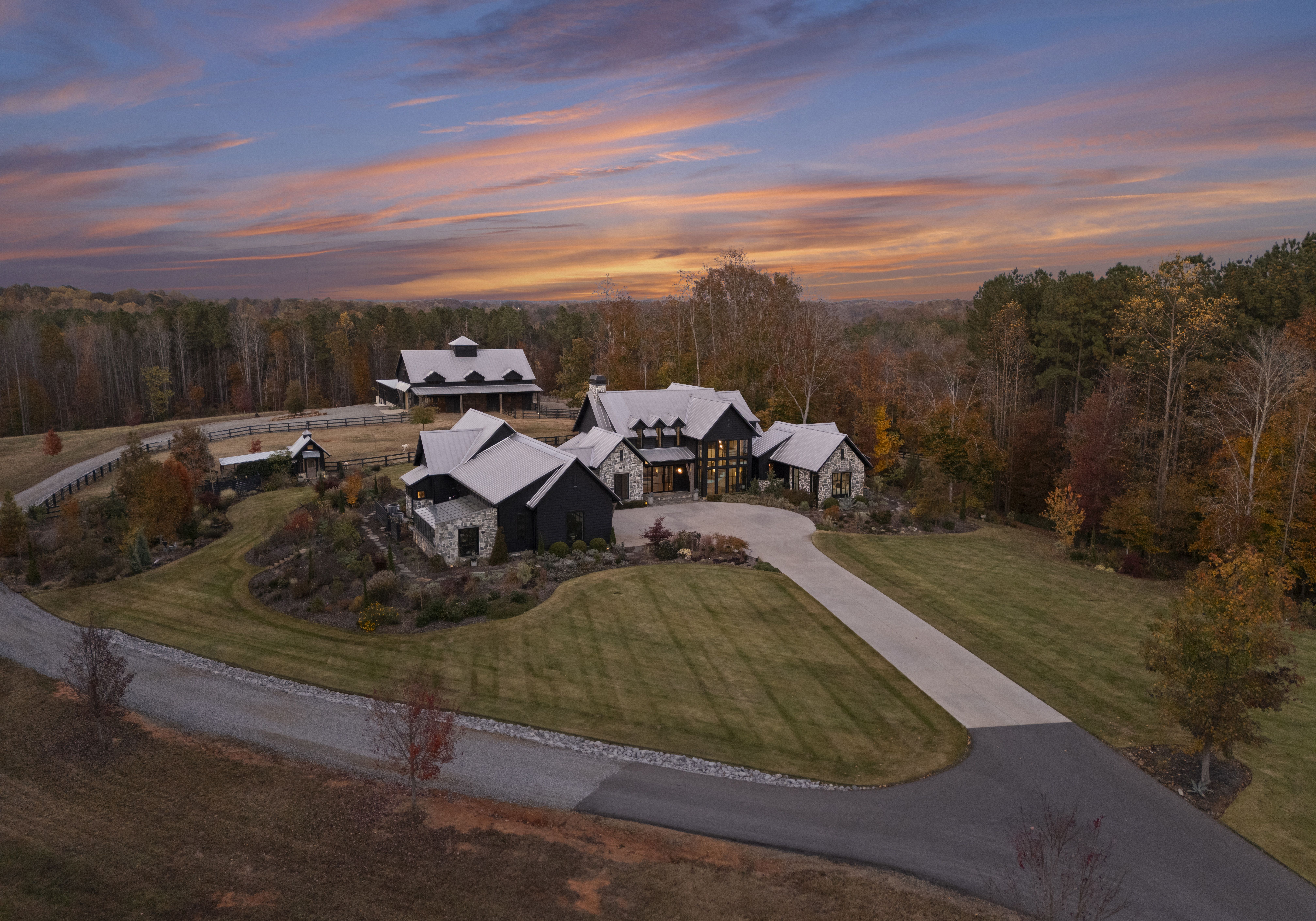 An aerial view of a large, modern stone house with multiple gabled roofs, dark siding, and a curved concrete driveway; autumn trees and a wooded landscape under a vibrant orange-pink sunset.