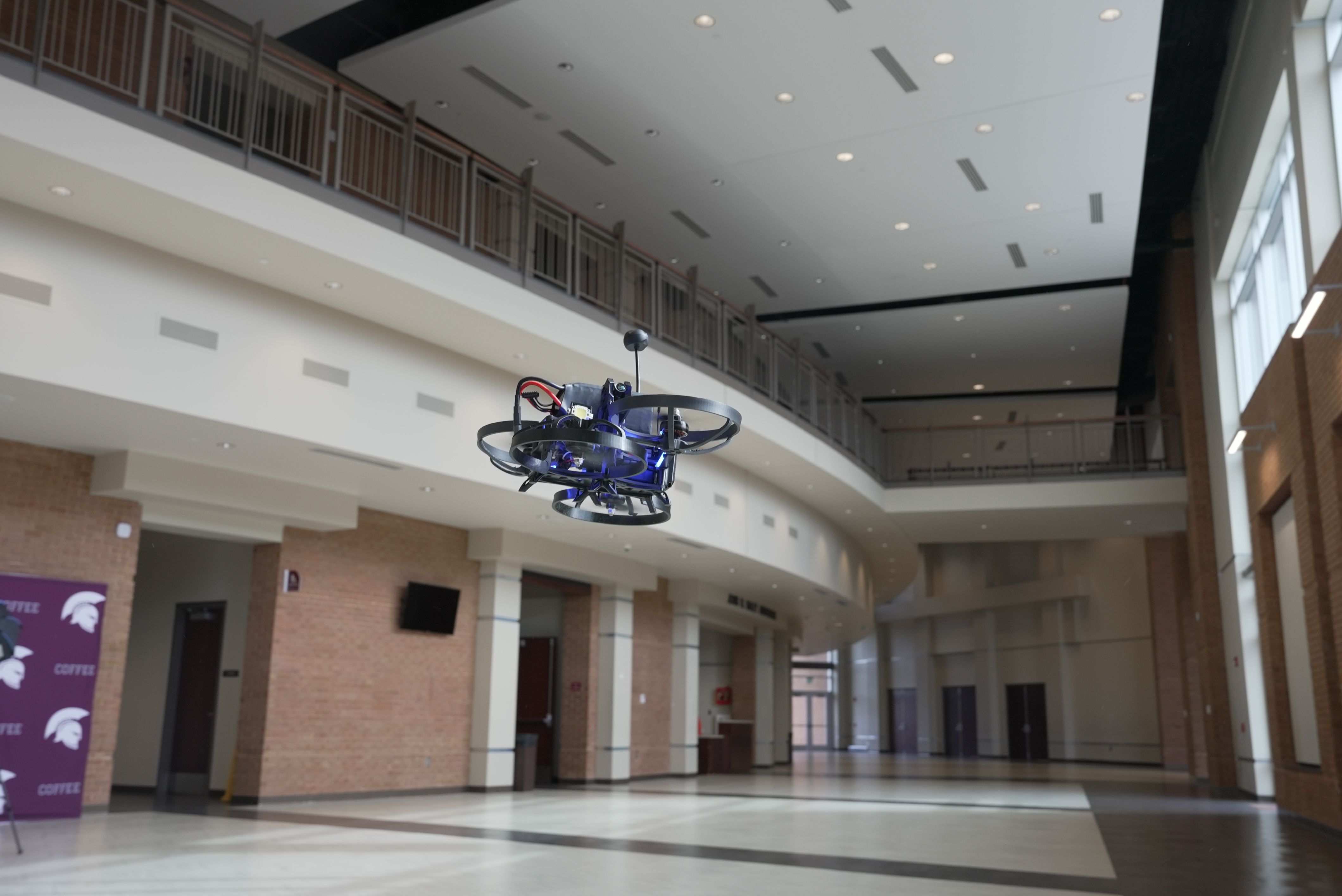 Blue quadcopter drone hovers in a spacious indoor atrium with brick walls, white columns, and an upper walkway railing; bright overhead lights and large windows line the right side.