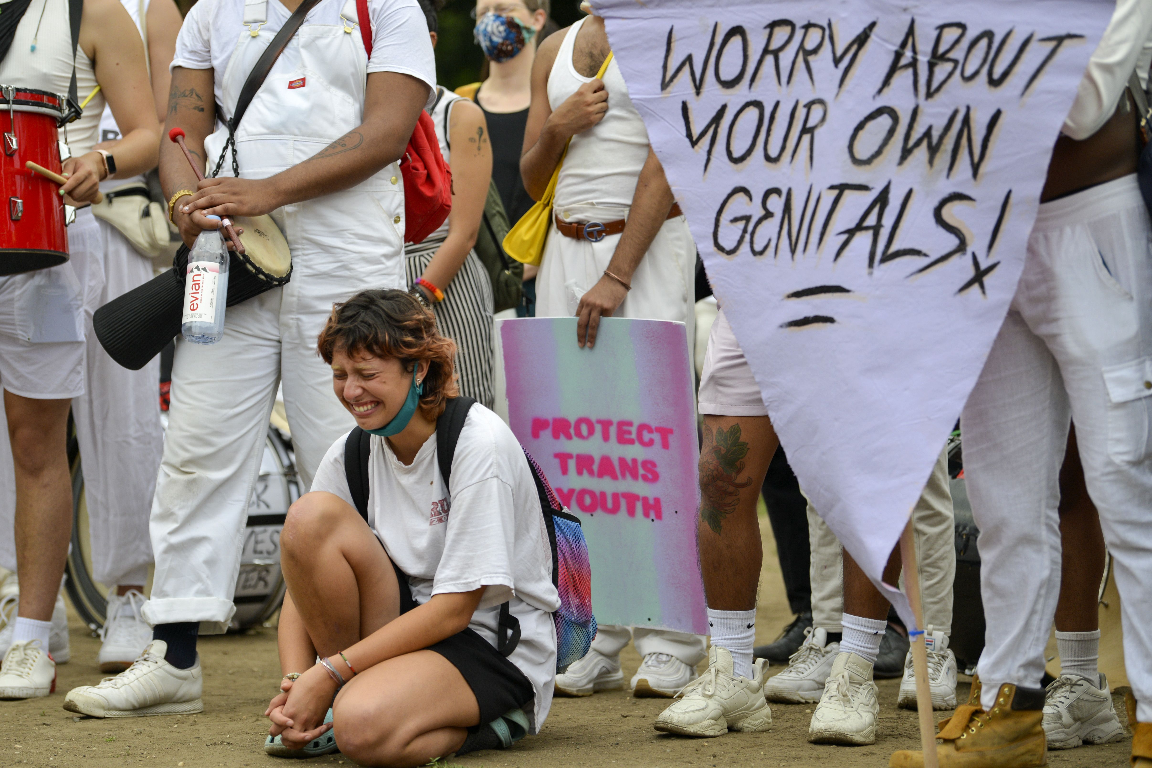 Photo of a person visibly emotional and kneeling on the ground with protesters holding signs behind them, one says "Worry about your own genitals"