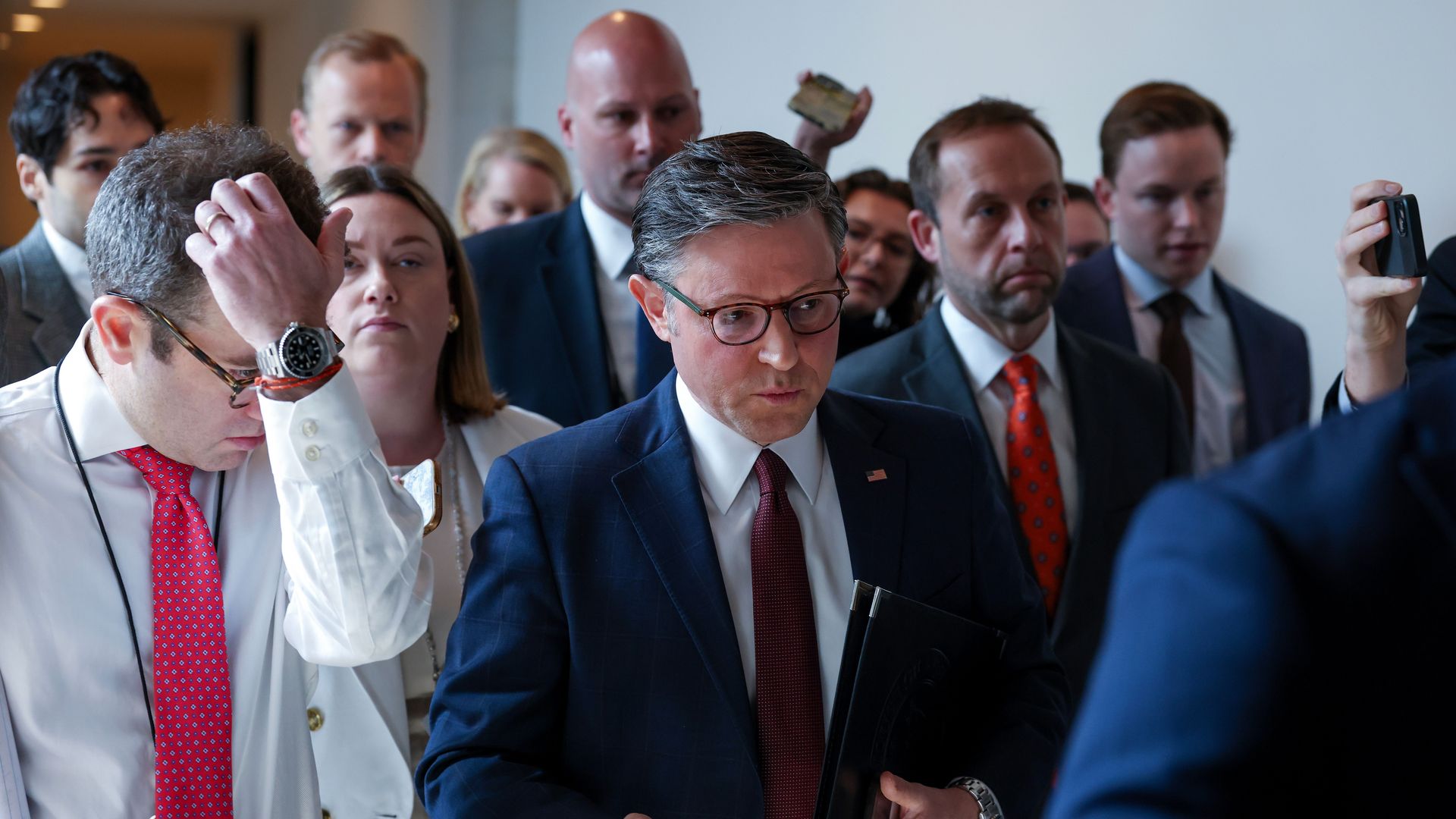 Man in a navy suit and red tie with glasses walks holding a black folder, surrounded by suited colleagues; a man on the left adjusts his hair while someone on the right records with a phone.