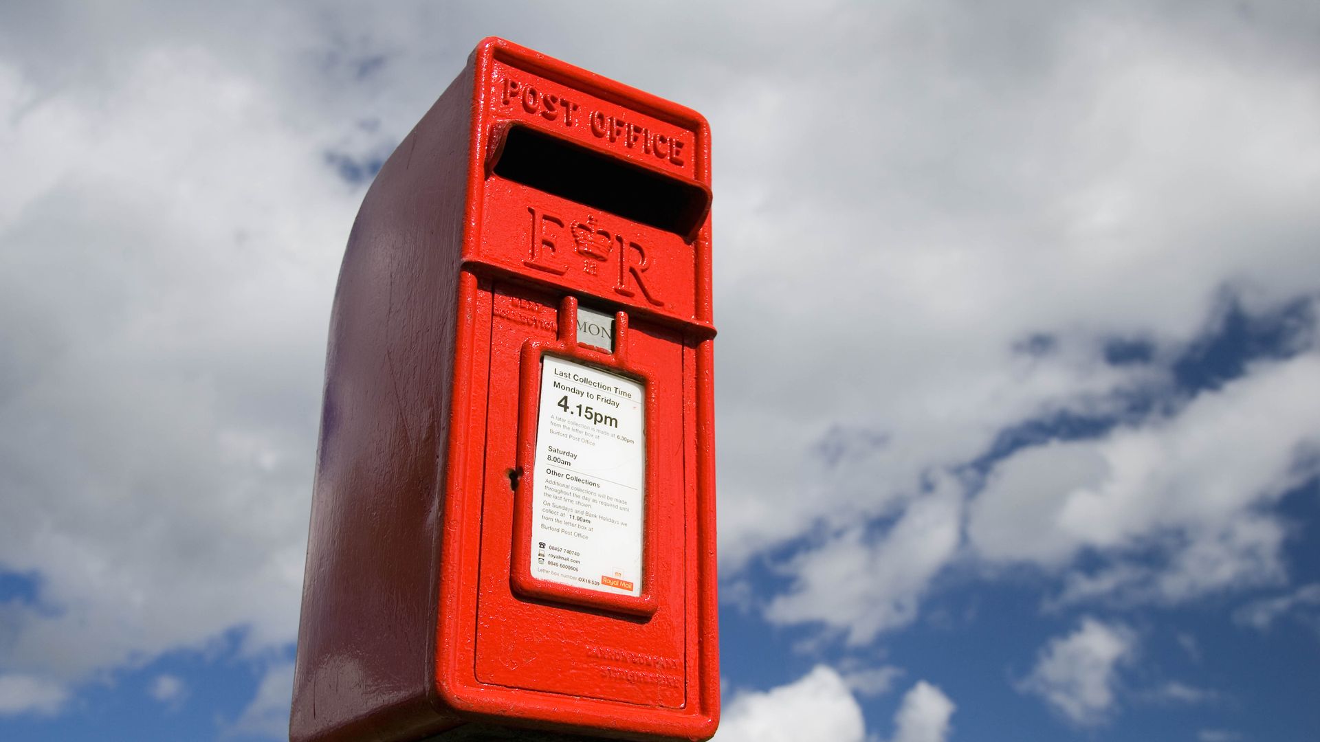 Bright red British post box with "Post Office" and "E II R" embossed, showing last collection time, set against a partly cloudy blue sky background.