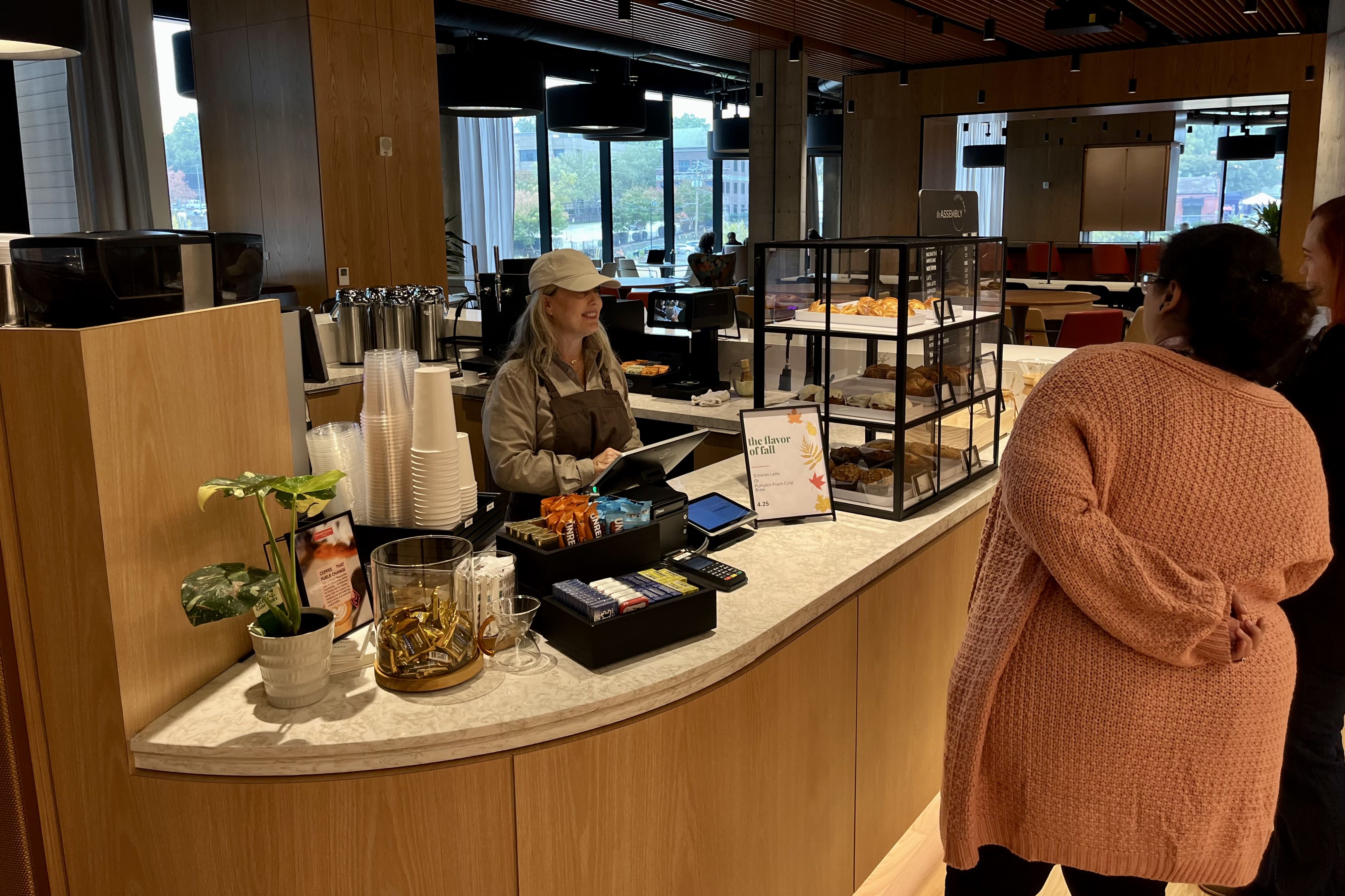 A woman in a beige cap and apron stands behind a counter with pastries and a sign reading "the flavor of fall" while two customers stand nearby in a modern cafe with wood accents and large windows.