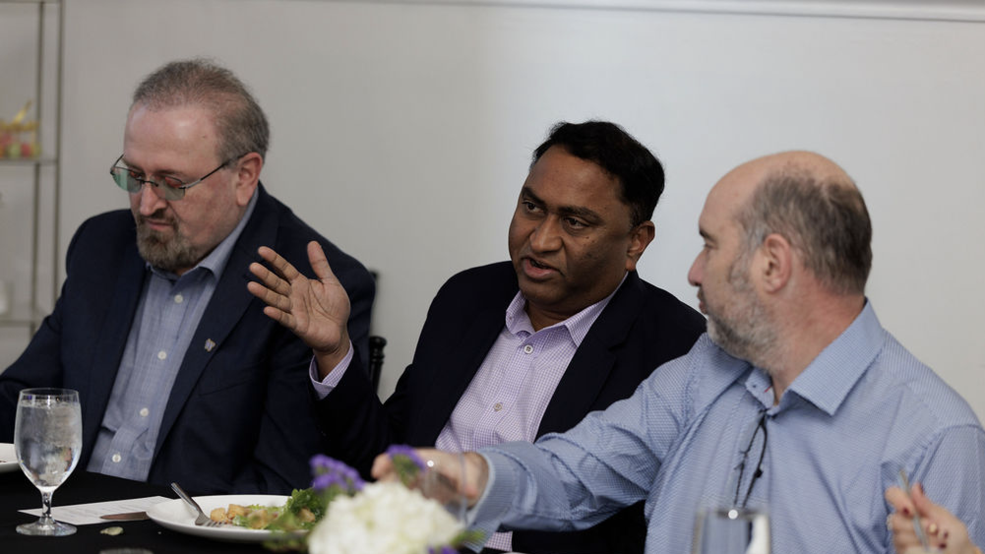 Three men in business attire sitting at a table with plates of food and glasses of water, engaged in conversation in a meeting or dining setting with a flower centerpiece.