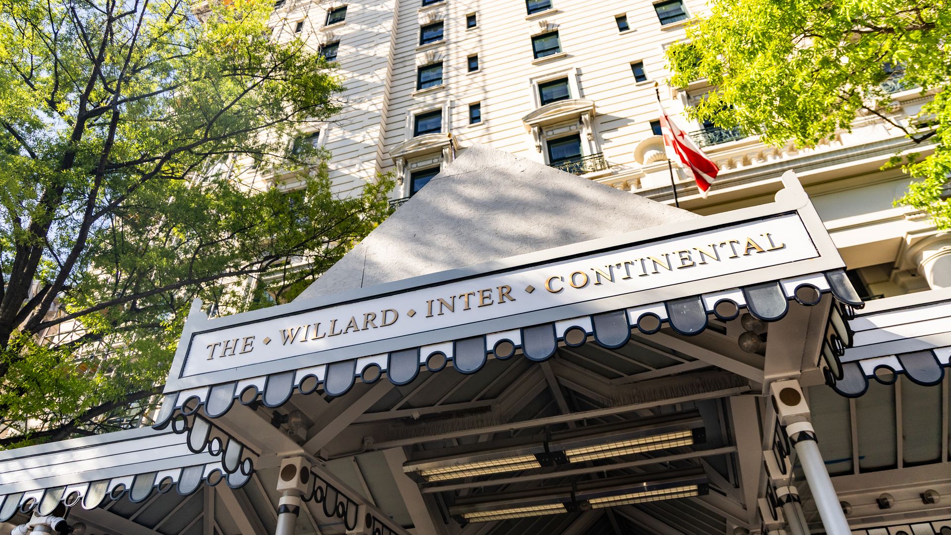 Cream-colored ornate hotel with tall windows and cornices; scaffolding on the roof, green trees framing the facade, a red-and-white flag at the entrance, and the marquee "The Willard InterContinental".