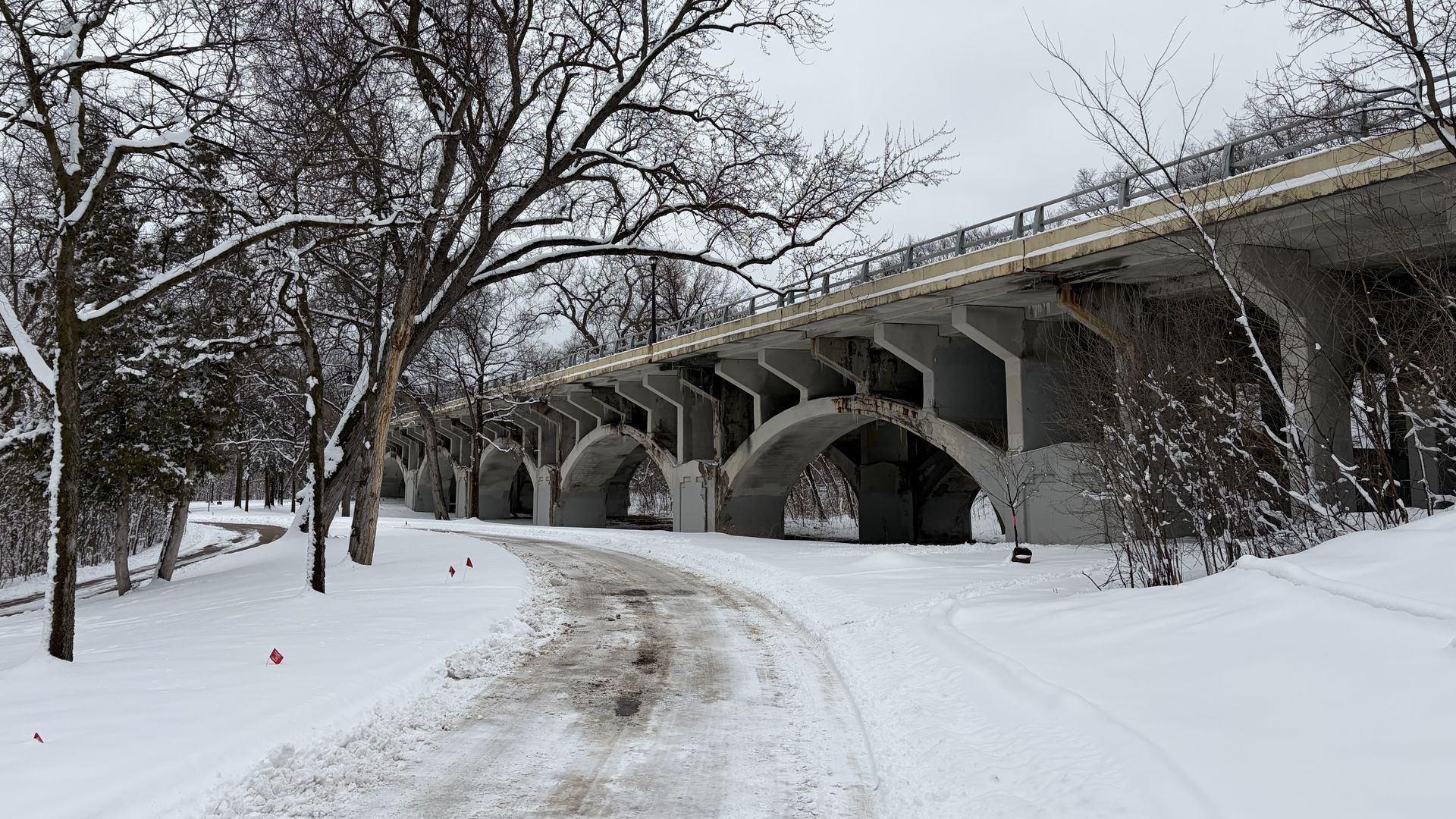 Snow-covered path curving under a large concrete bridge with arch supports, surrounded by bare trees coated with snow on an overcast winter day.