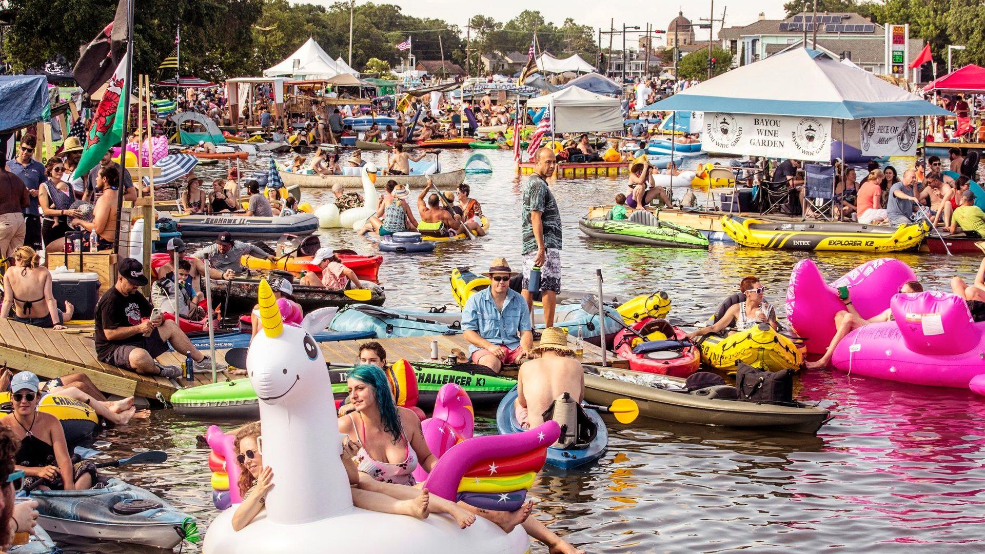 Crowded lively water festival with people in colorful kayaks and inflatable floats shaped like unicorns and flamingos enjoying sunny weather on calm water.
