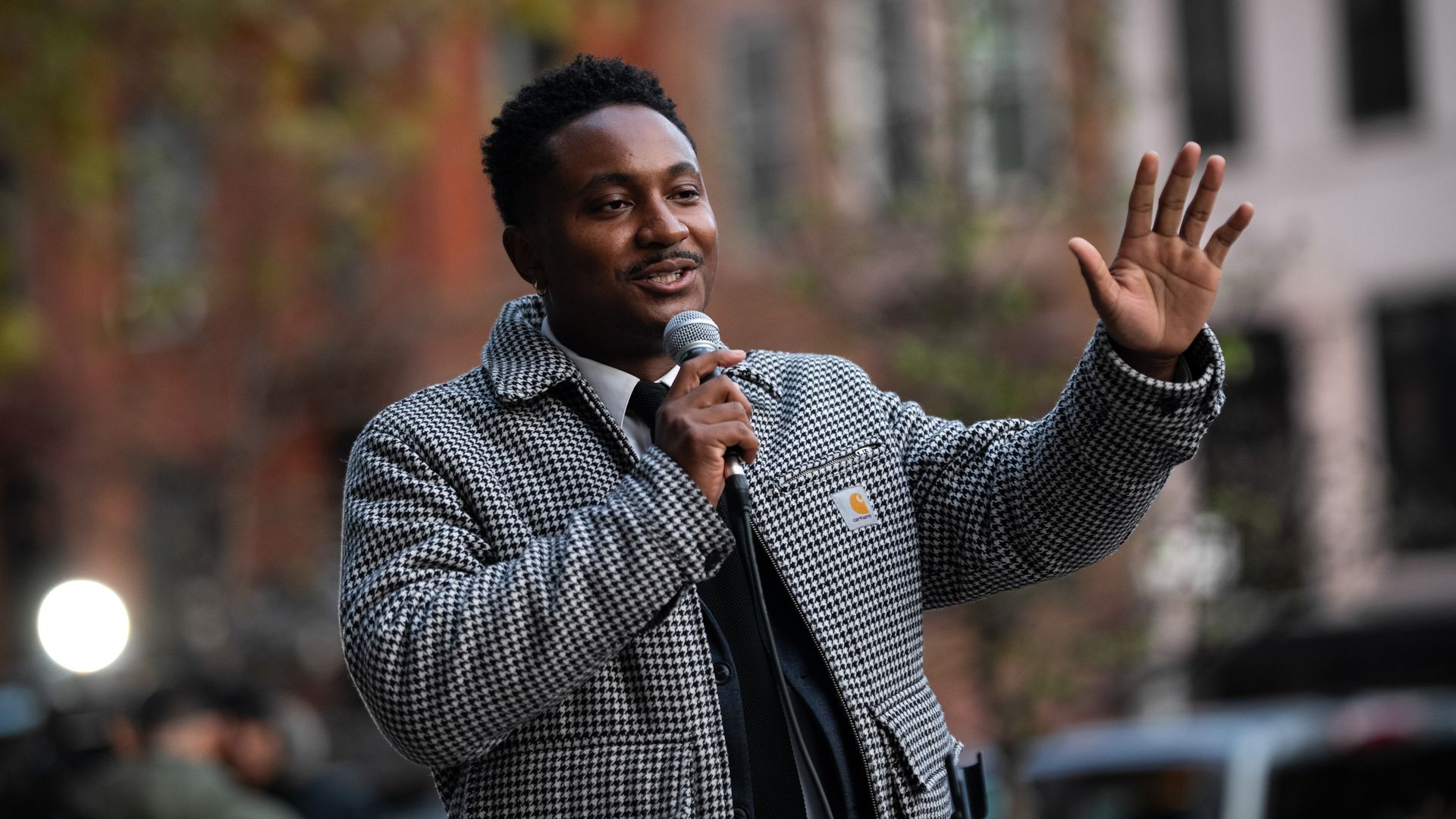 Chi Osse, holding a microphone in one hand and holding up his other hand while wearing a white and black jacket and standing in front of townhouses.