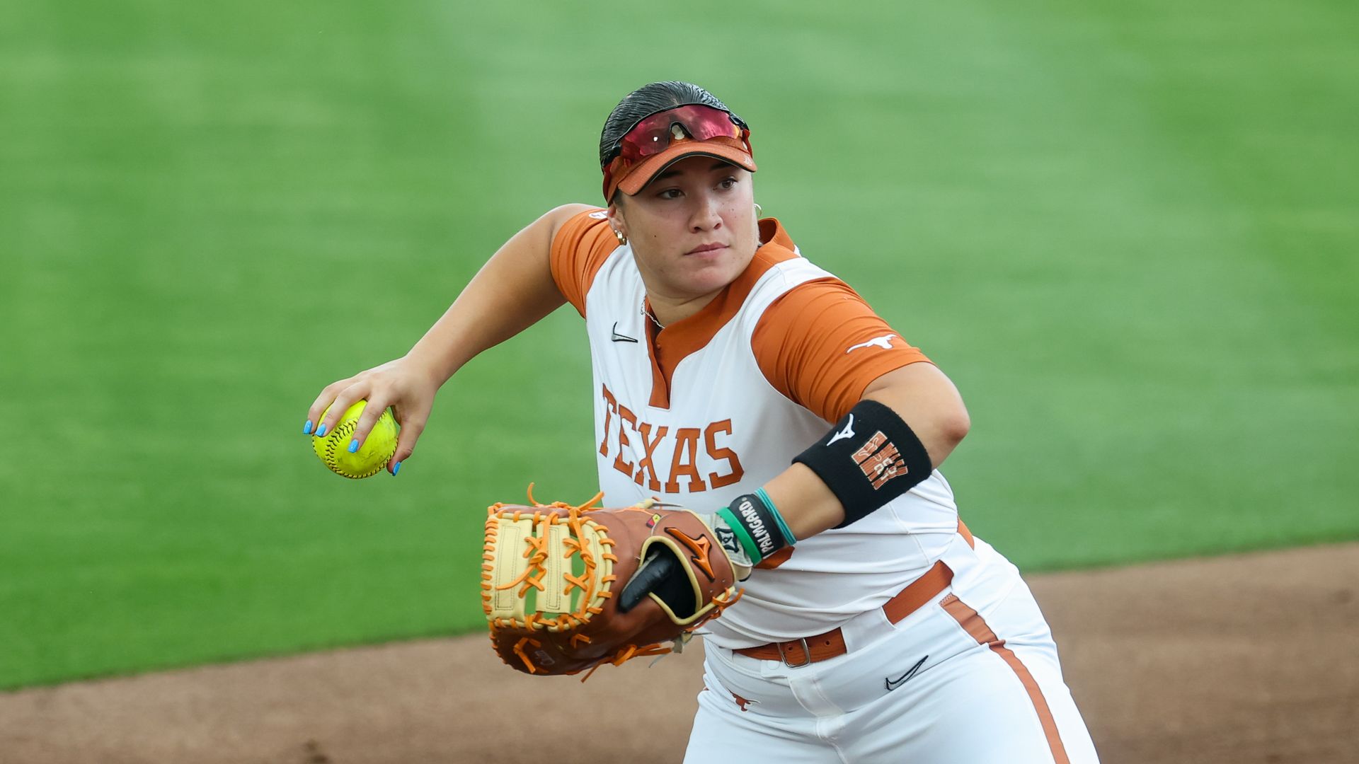 Texas softball player in orange and white uniform, orange cap with sunglasses, glove on left hand and bright yellow ball in hand, on a dirt infield with green grass in the background.