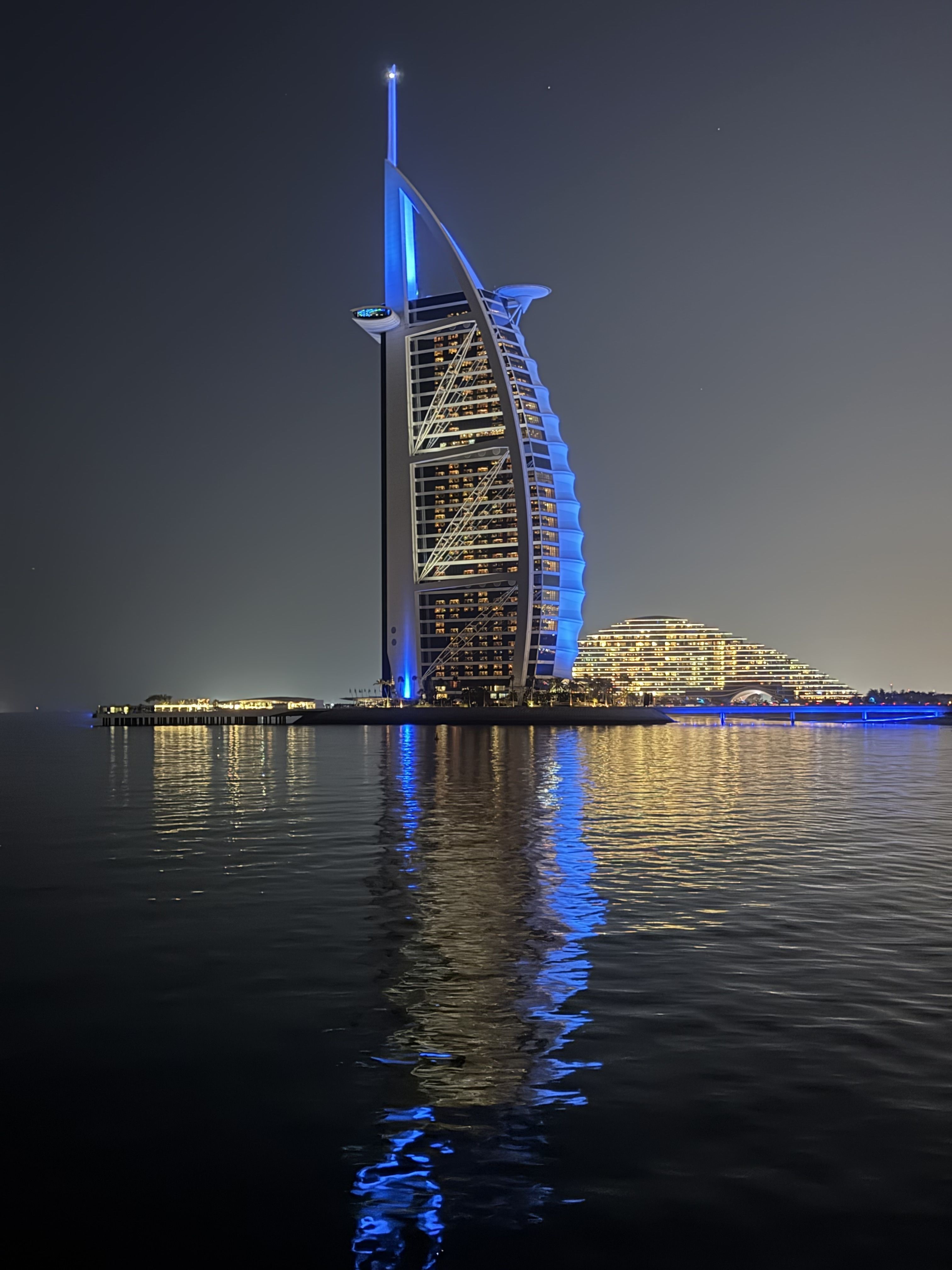 Burj Al Arab hotel lit with blue and white lights at night, reflecting on calm water, with a lit, dome-shaped building in the background under a dark sky.