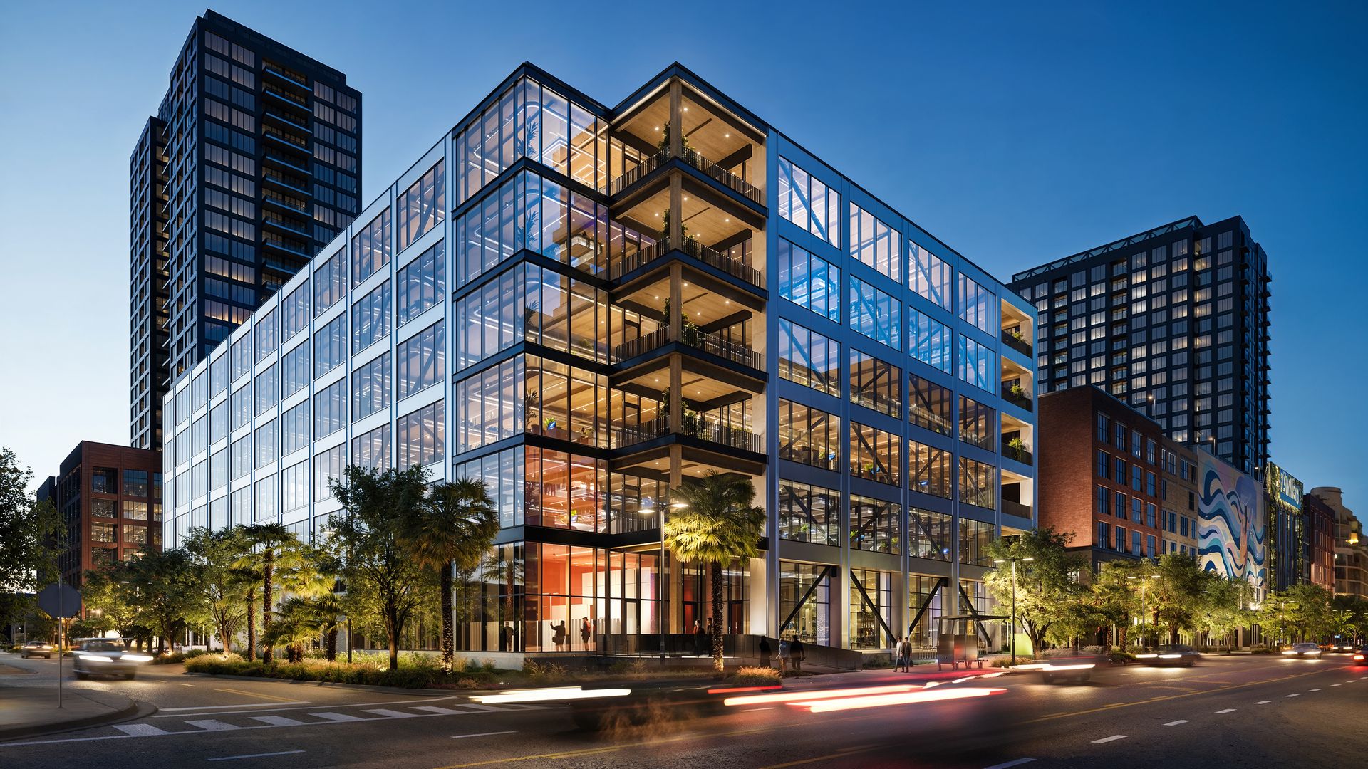 Modern glass office complex at dusk with illuminated interiors, glass facade, and balconies; palm trees line the street as light trails sweep the road.