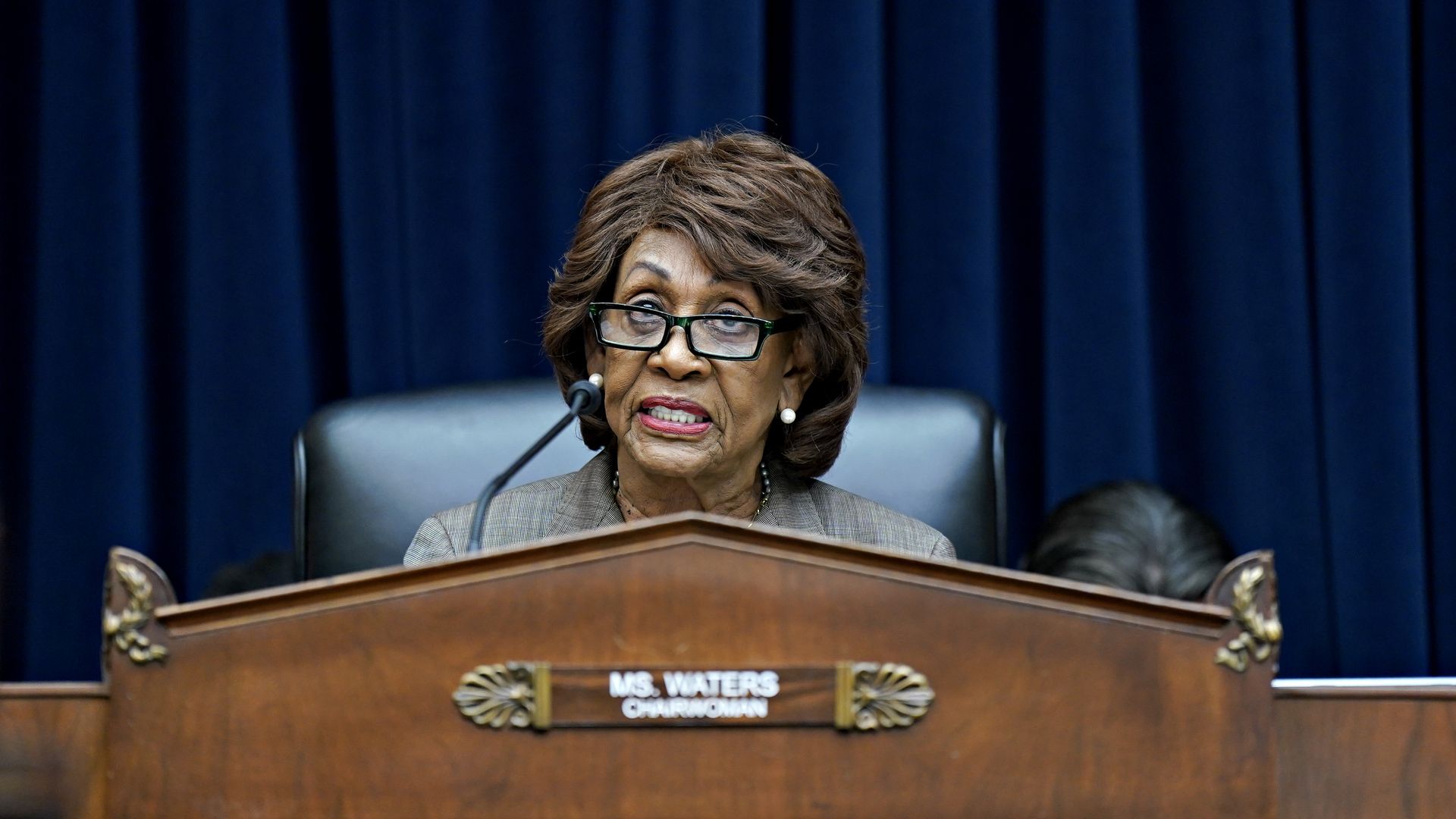 Maxine Waters, wearing glasses and a gray suit behind a committee dais.