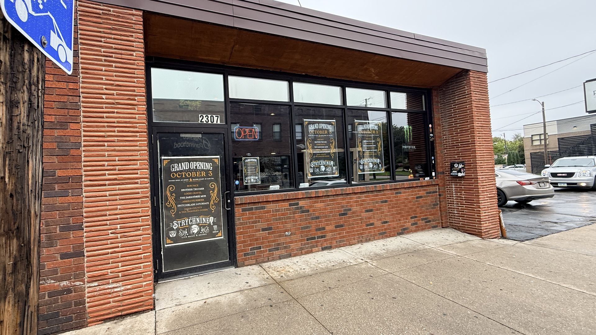 Brick storefront with large black-framed windows, orange neon "OPEN" sign, and Grand Opening posters. Door shows 2307; blue handicap sign on the left; street with parked cars in the background.
