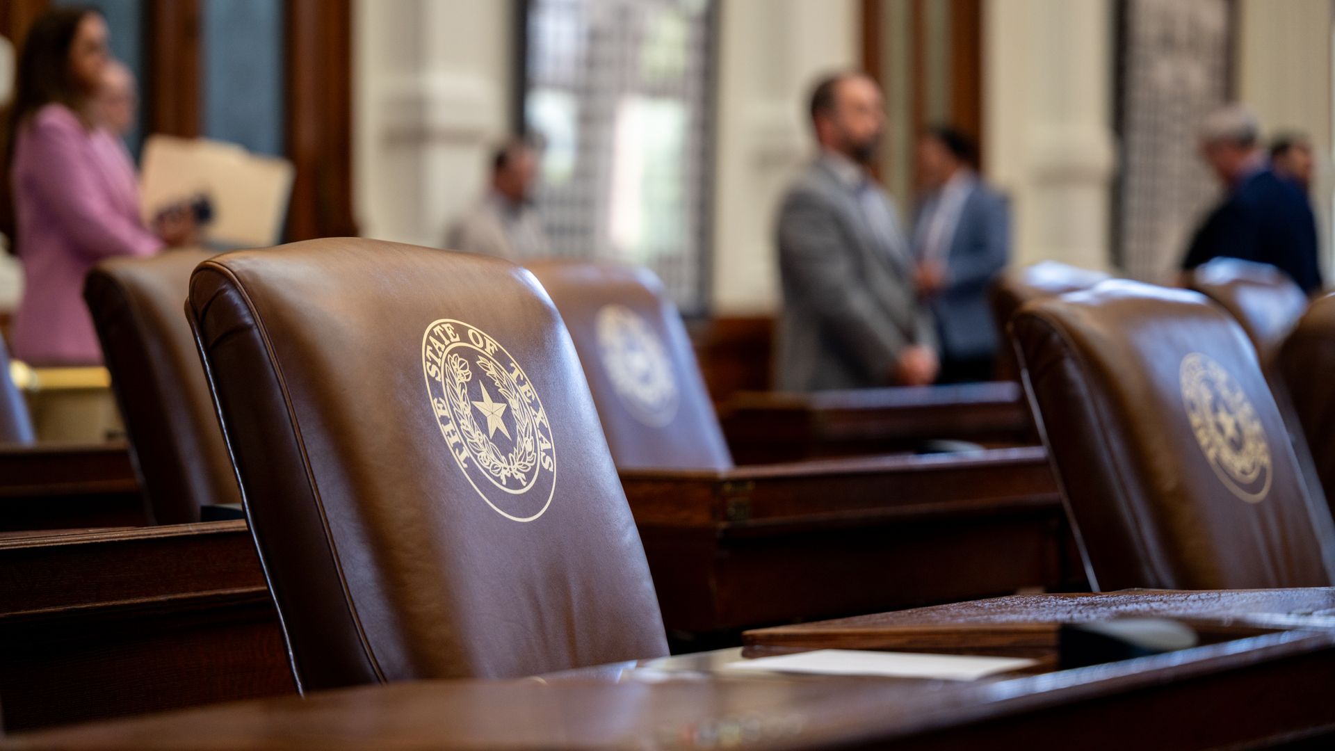 Brown leather chairs with "The State of Texas" seal in a legislative chamber, blurred background shows people standing and talking.