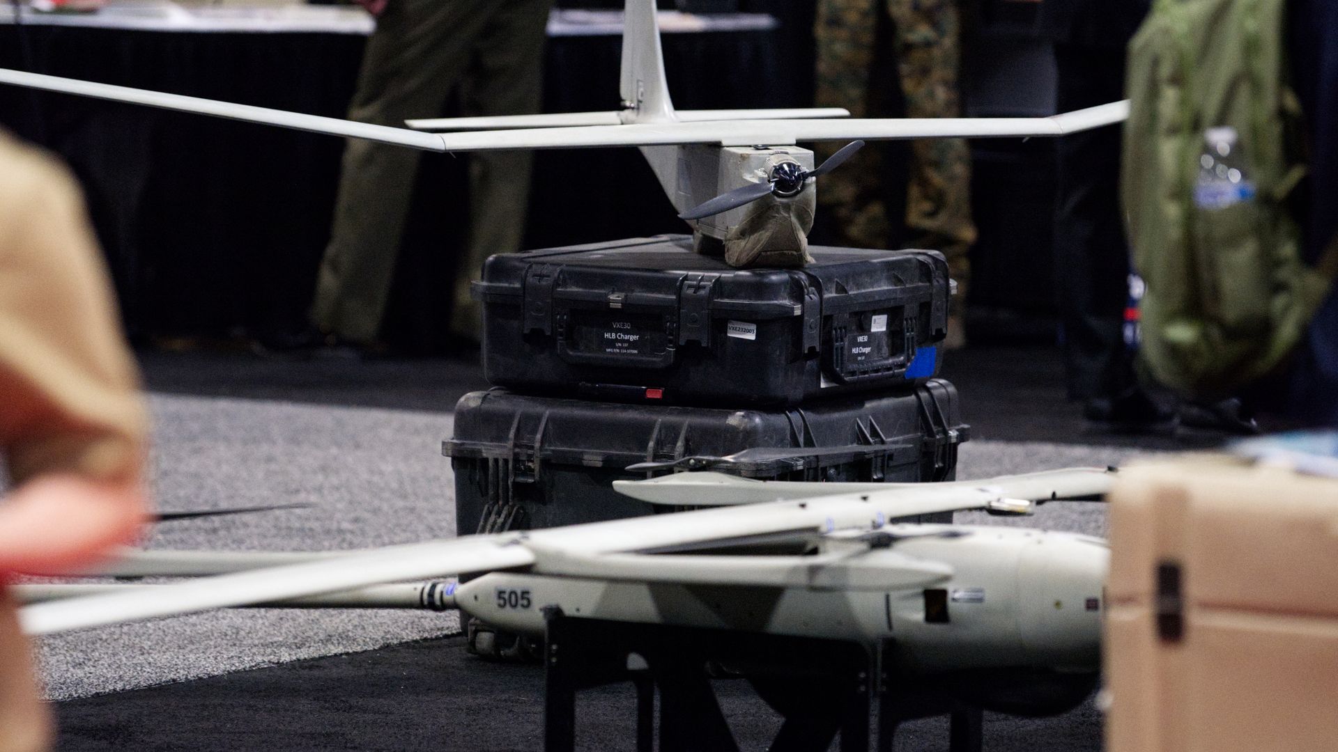 Two drones sit on the floor at a defense conference. People walk by, on the left and right sides of the photo.