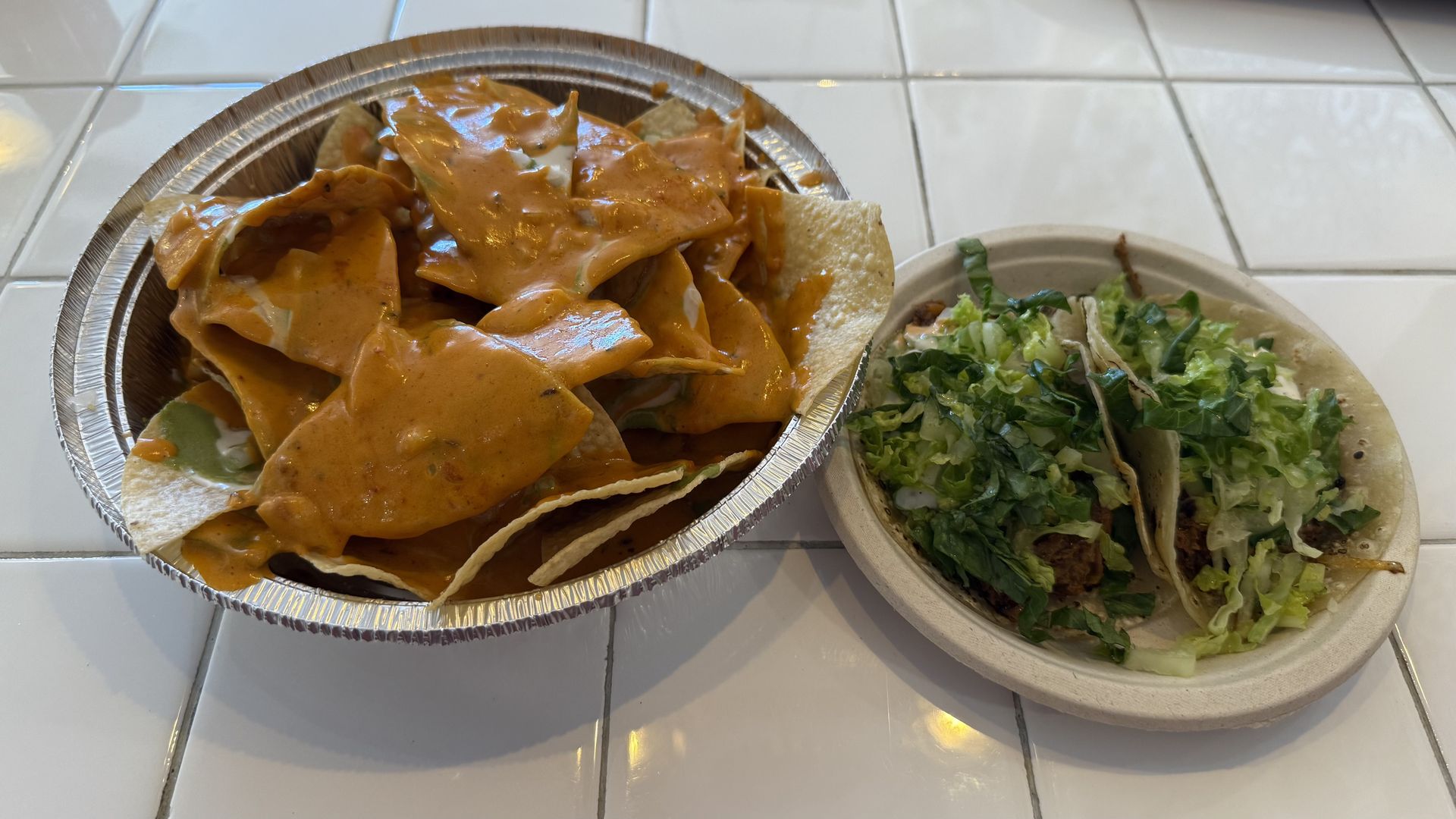 A metal take-out dish filled with tortilla chips covered in an orange-brown sauce, next to a paper plate with two lamb tacos.