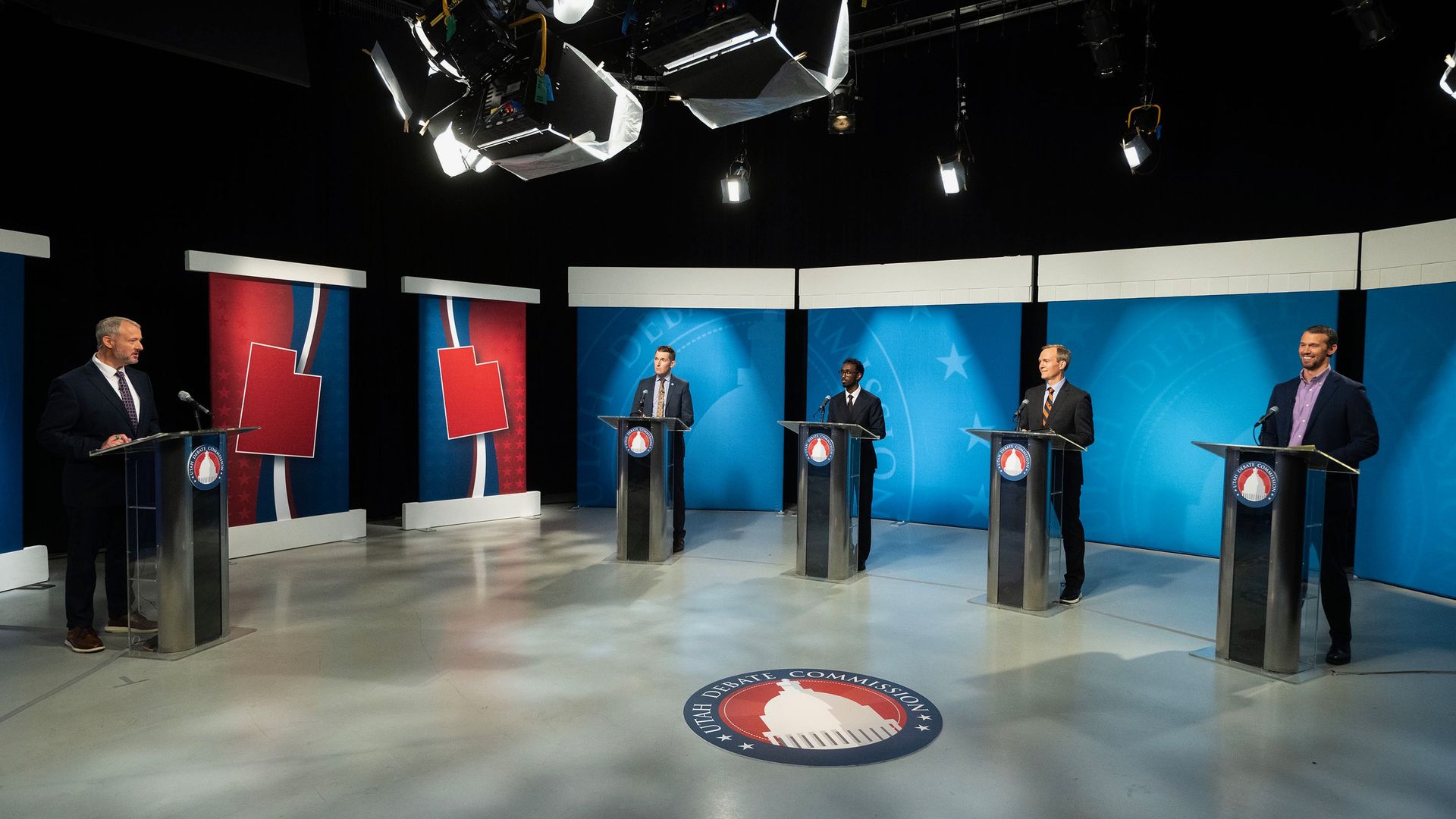 Five speakers stand behind podiums on a TV debate stage with red and blue backdrops, bright overhead lights, and a circular debate commission logo on the floor.