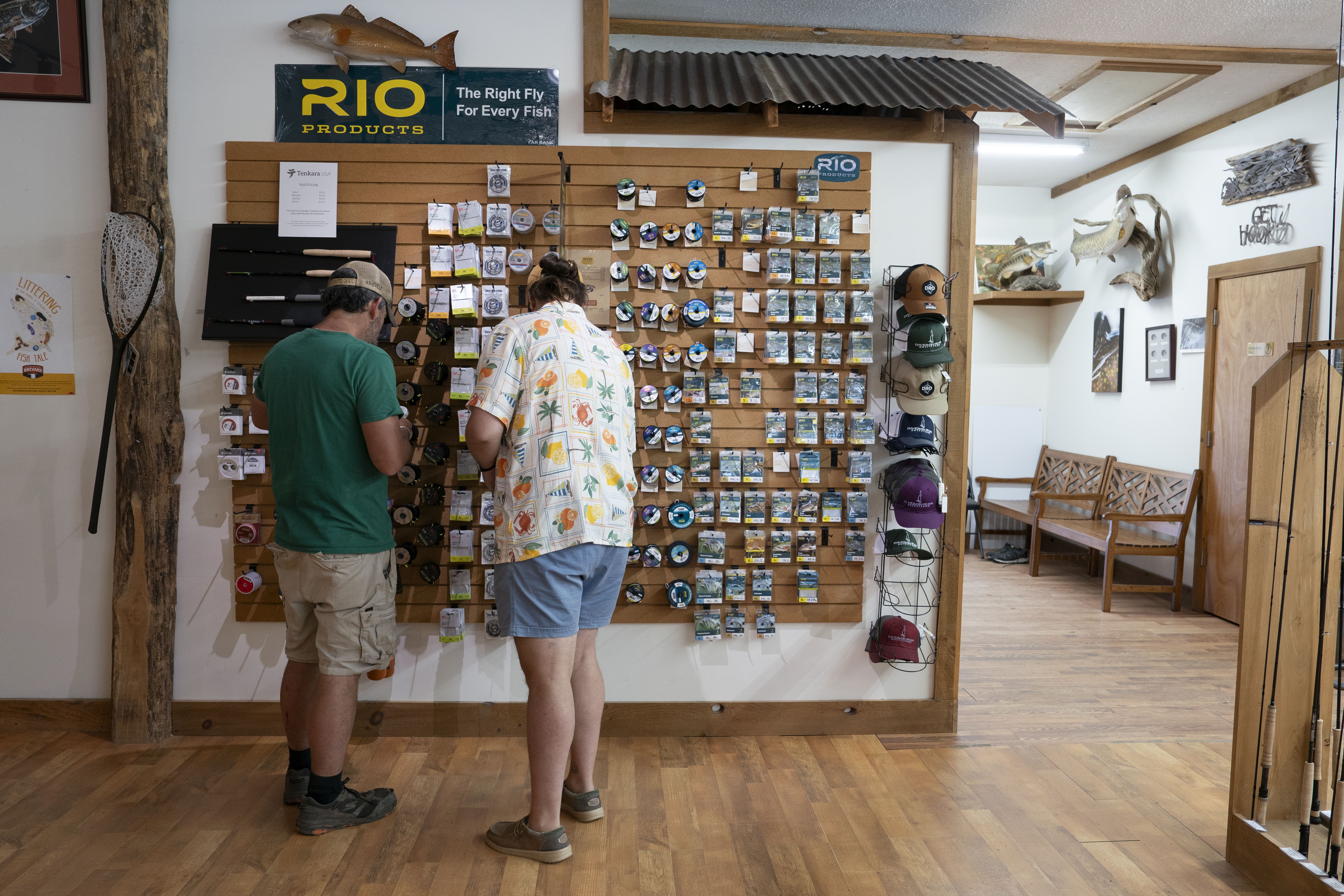Two people browse fishing gear inside a shop, looking closely at a wall display of fly-fishing products, reels, and accessories. Hats and rods are also displayed.