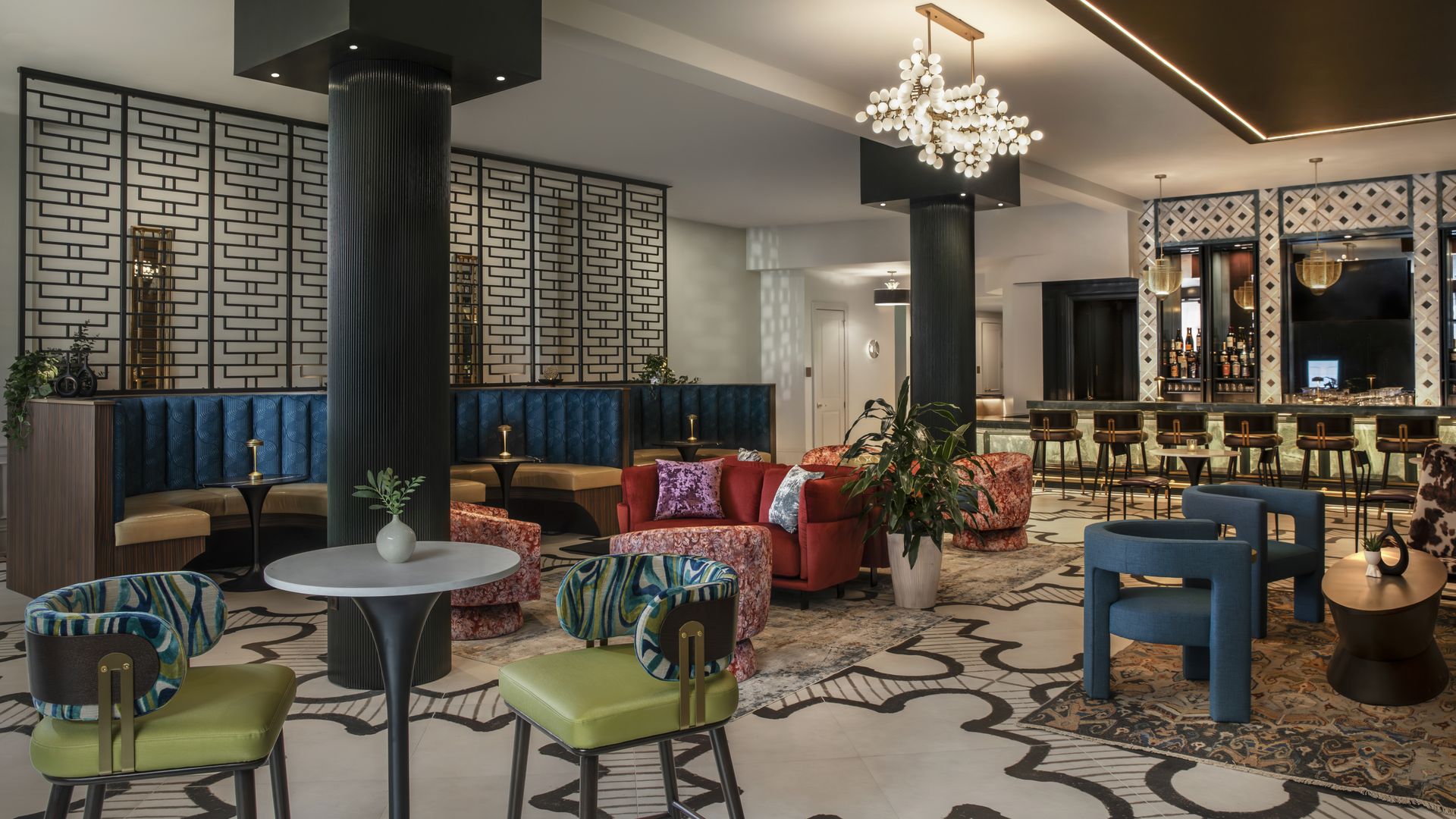 Upscale hotel lounge with a tall dark column and decorative screens on the left. Blue tufted banquettes, bright colorful chairs, round tables, potted plants, and a well-lit bar area in back.