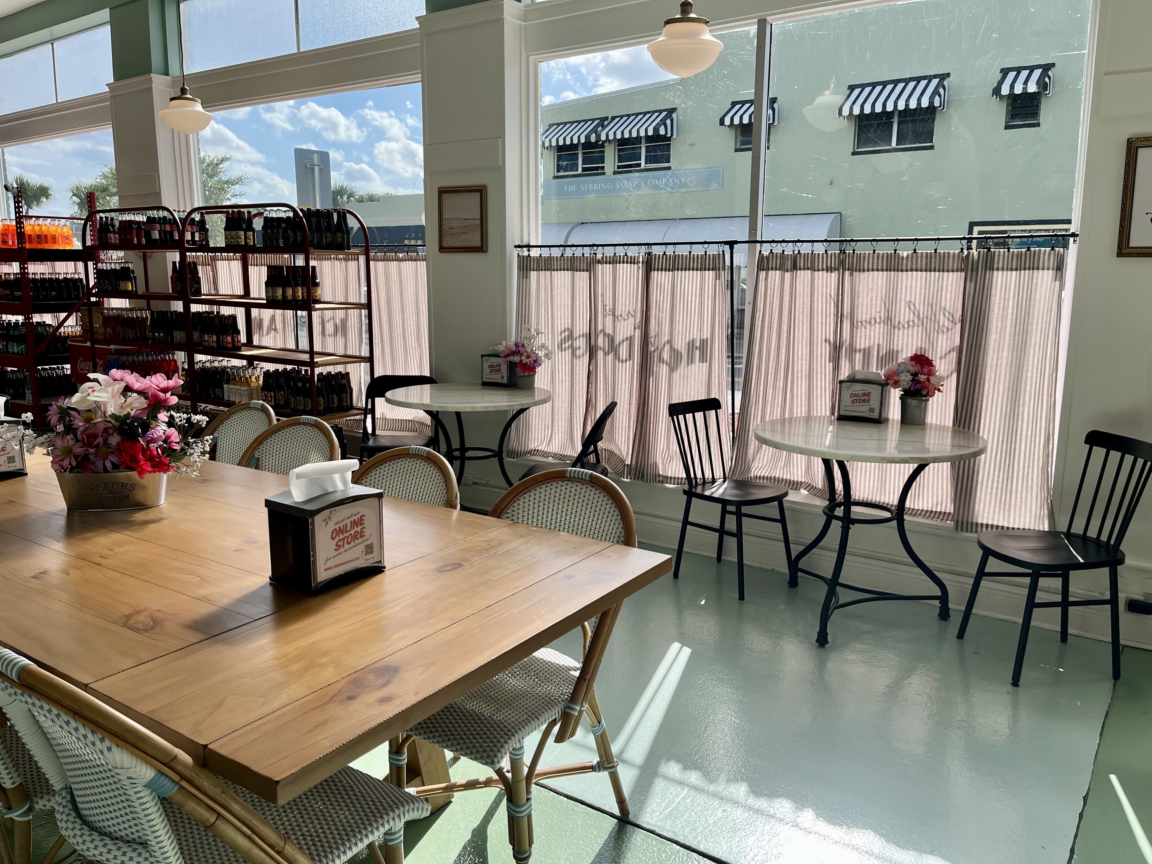 The brightly lit interior of a shop with shelves of bottled soda, striped curtains on the windows, and tables and chairs.