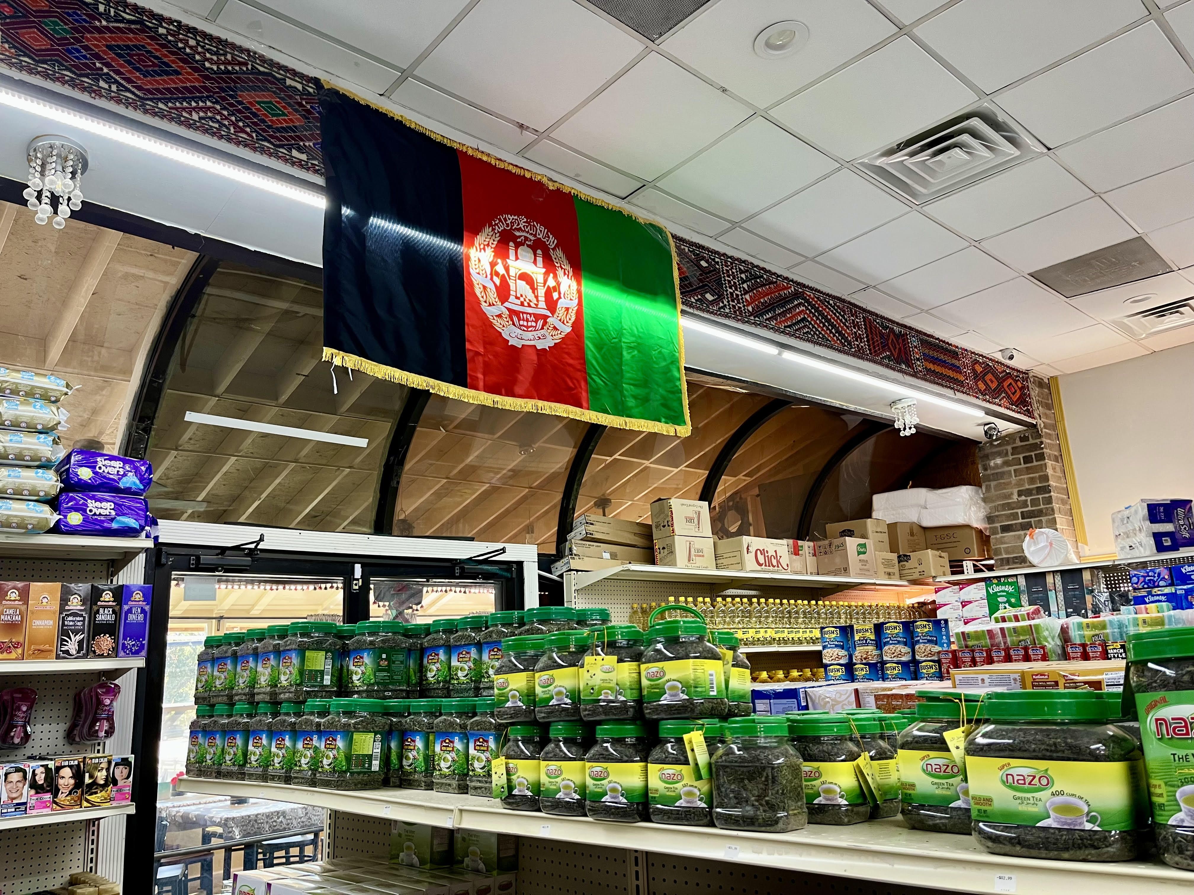 An Afghanistan flag hangs above store shelves stocked with green tea jars, various food items, and household products inside Kabul International Food Market and Restaurant in San Antonio.