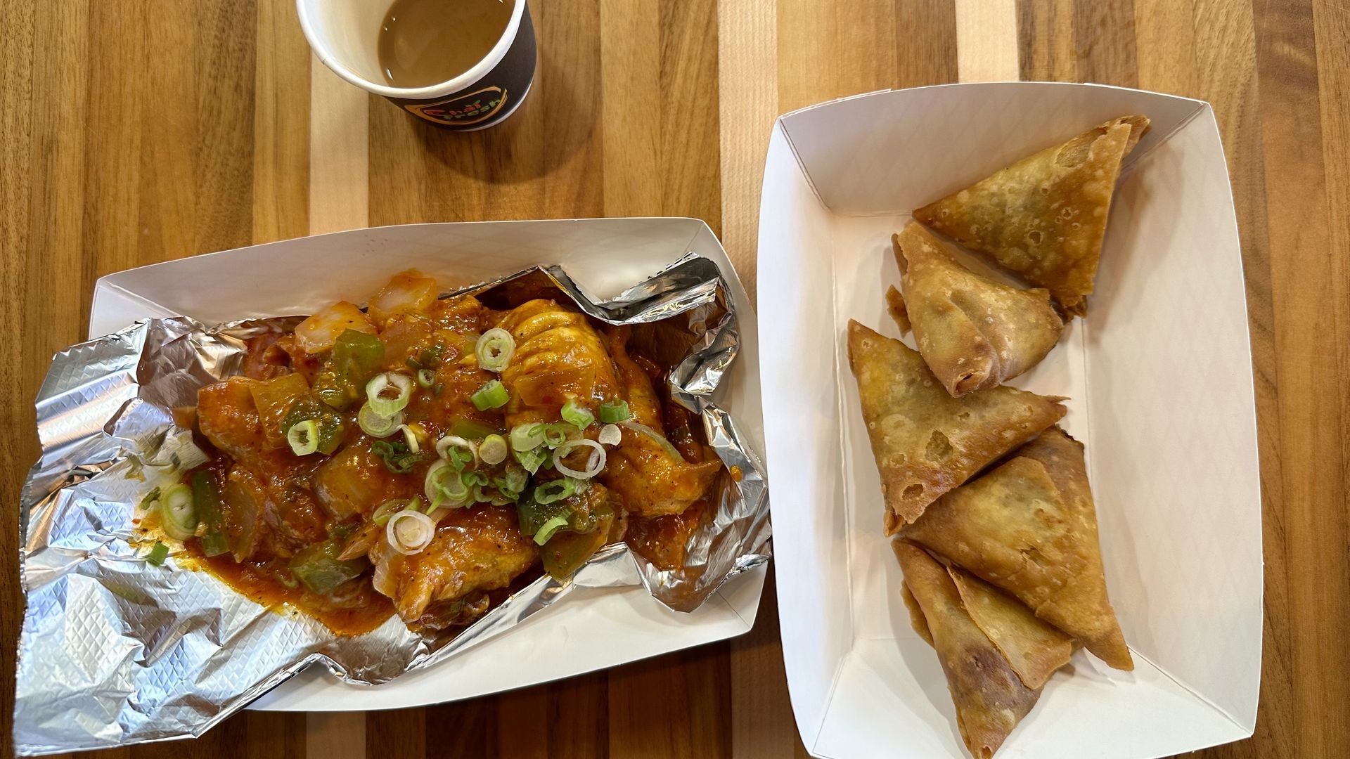 Top-down view of a wooden table with takeout: a cup of coffee or tea, a foil-wrapped spicy dish topped with green onions, and a white tray of triangular fried pastries.